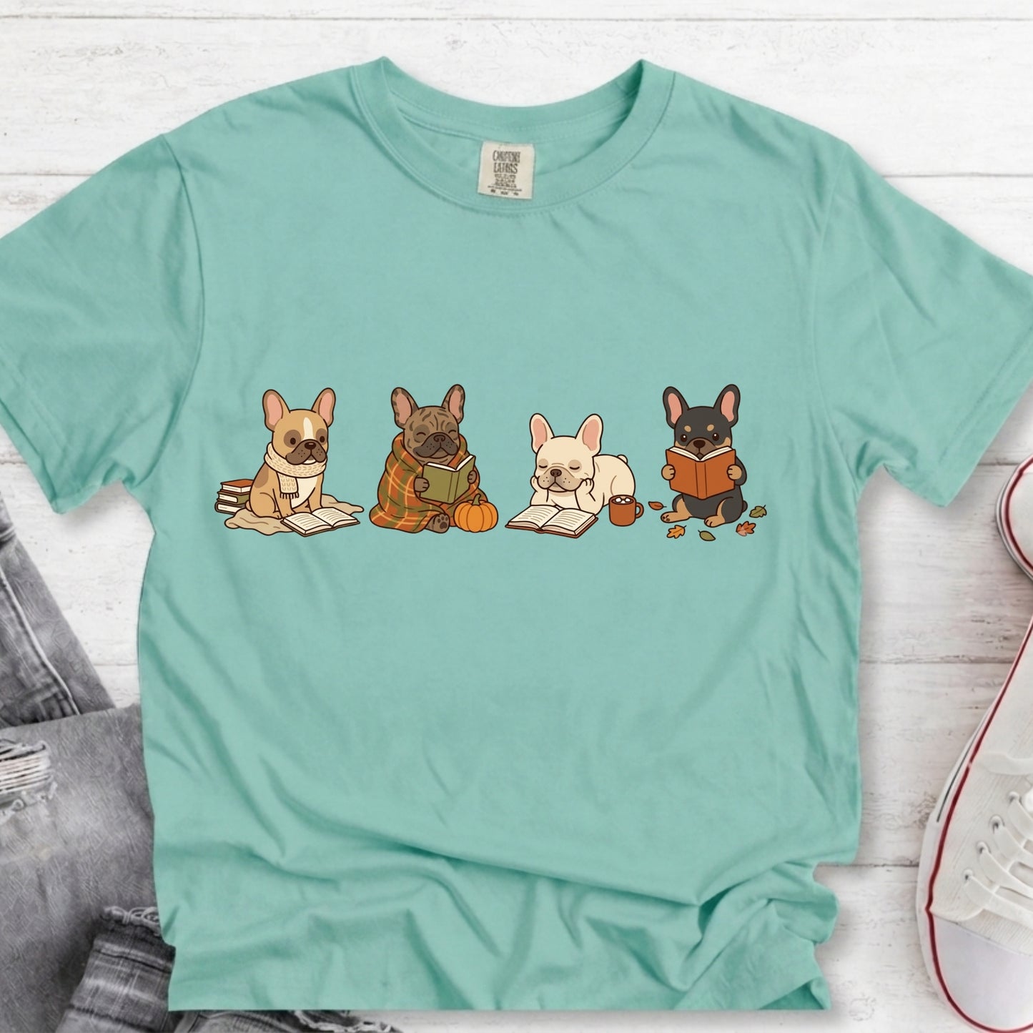 A mint green short-sleeve t-shirt with a graphic of four French Bulldogs reading books, laid flat on a white wood background with jeans and a sneaker partially visible.