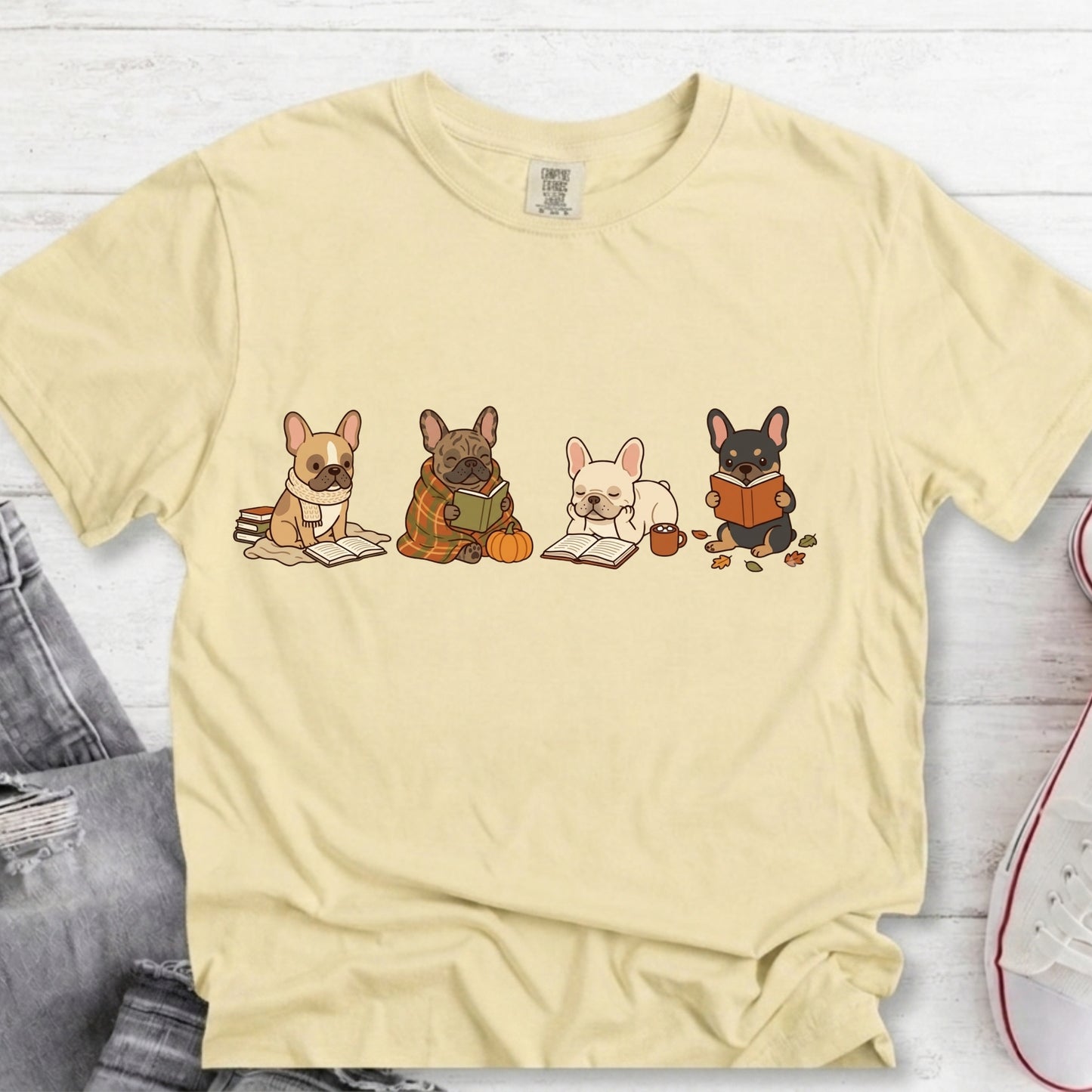 A pale yellow short-sleeve t-shirt with a graphic of four French Bulldogs reading books, styled in a flat-lay on a white wood background with jeans and a sneaker partially visible.