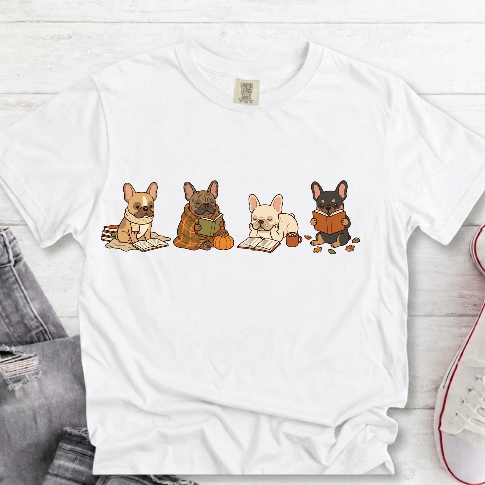 A white short-sleeve crewneck t-shirt featuring a graphic of four cute French Bulldog characters reading books, styled in a flat-lay arrangement on a white wood background with jeans and a sneaker partially visible.