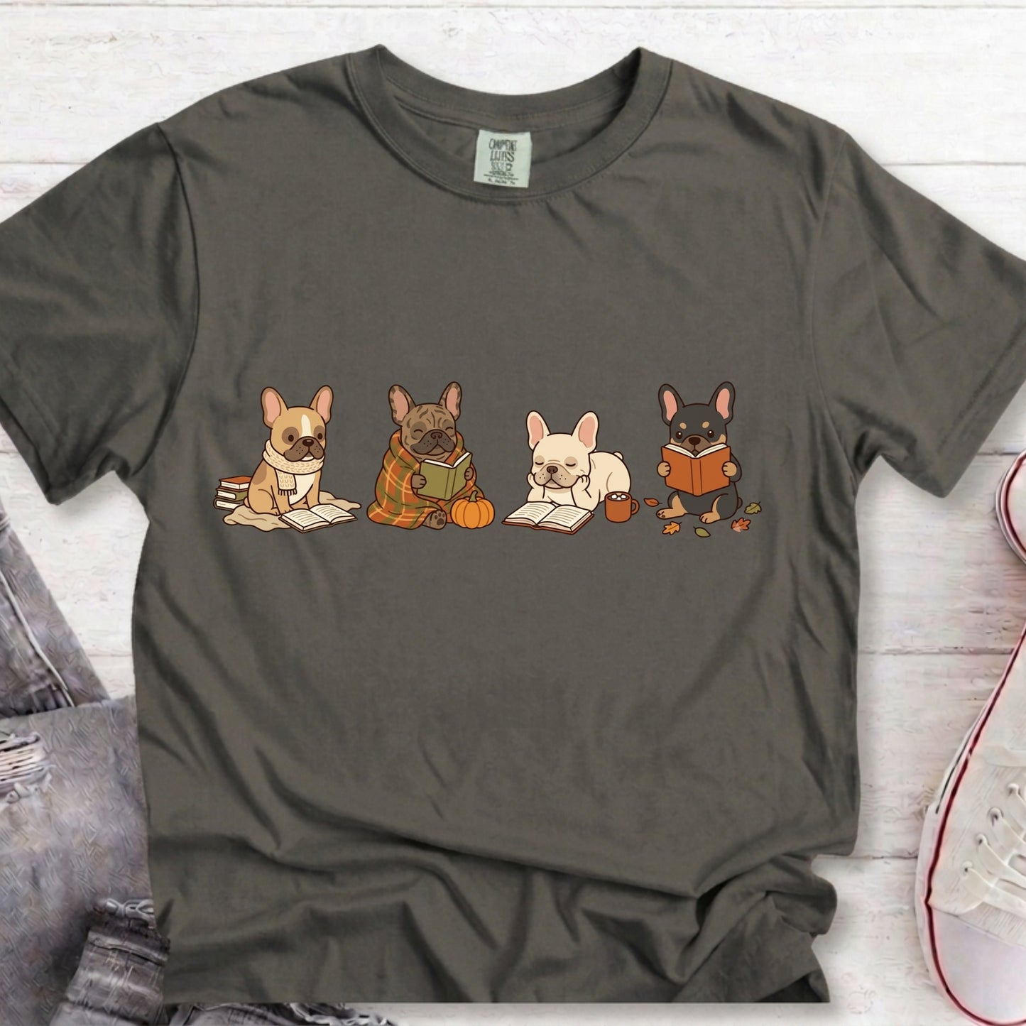 A dark charcoal gray t-shirt featuring a graphic of four cute French Bulldog characters reading books, styled in a flat-lay on a white wood background with jeans and a sneaker partially visible.