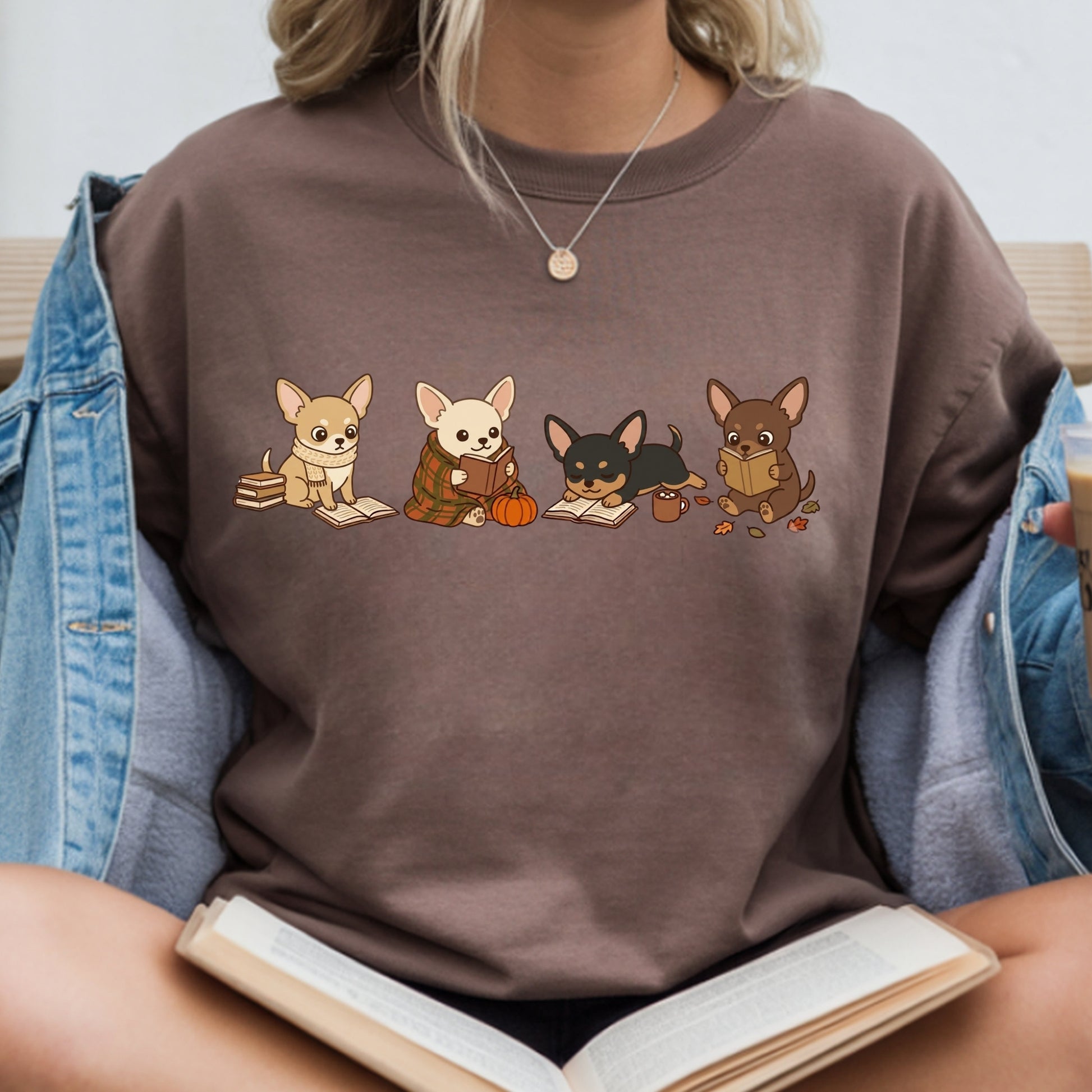 A person wearing a brown crewneck sweatshirt with a graphic of four cute cartoon Chihuahua dogs in autumn clothing, styled with an open denim jacket and holding an open book.