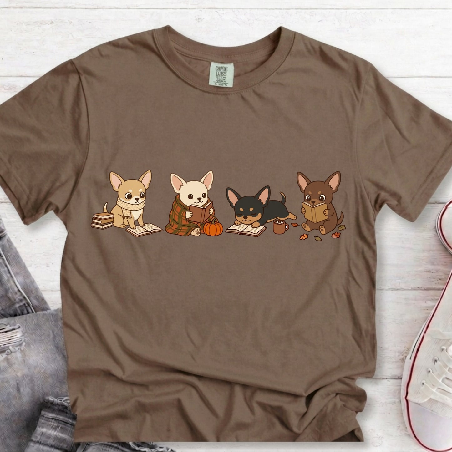A brown short-sleeve t-shirt laid flat on a white wood background, featuring a graphic of four cute cartoon Chihuahua dogs sitting with books and a pumpkin. A pair of gray jeans and a white sneaker are partially visible as styling props.