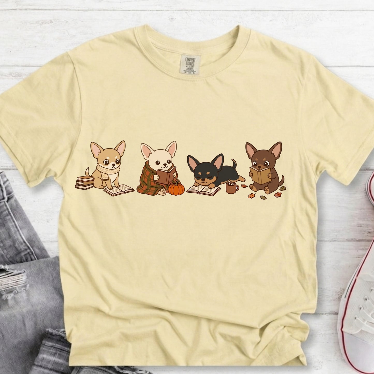 A pale yellow short-sleeve t-shirt with a graphic of four cute cartoon Chihuahua dogs reading, styled in a flat-lay on a white wood background with jeans and a sneaker partially visible.
