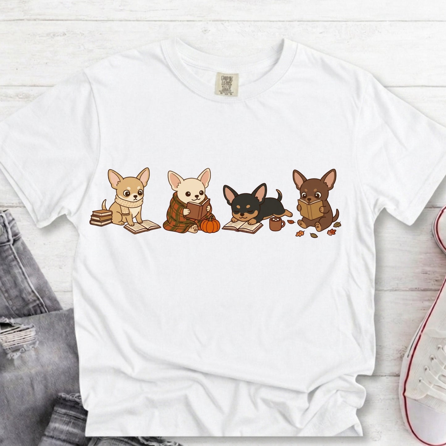 A white short-sleeve crewneck t-shirt featuring a graphic print of four cute cartoon Chihuahua dogs reading books, styled in a flat-lay on a white wood background with jeans and a sneaker partially visible.