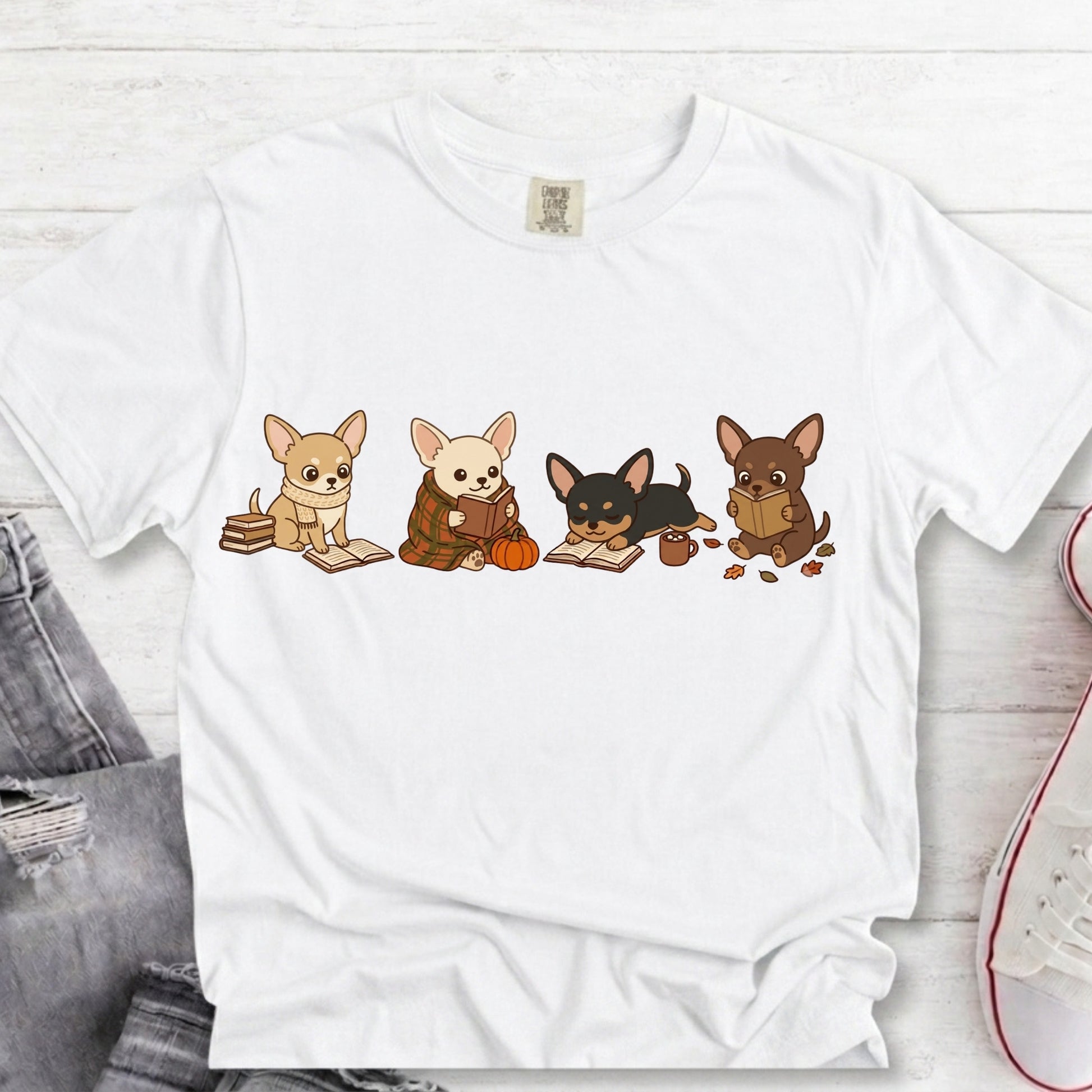 A white short-sleeve crewneck t-shirt featuring a graphic print of four cute cartoon Chihuahua dogs reading books, styled in a flat-lay on a white wood background with jeans and a sneaker partially visible.
