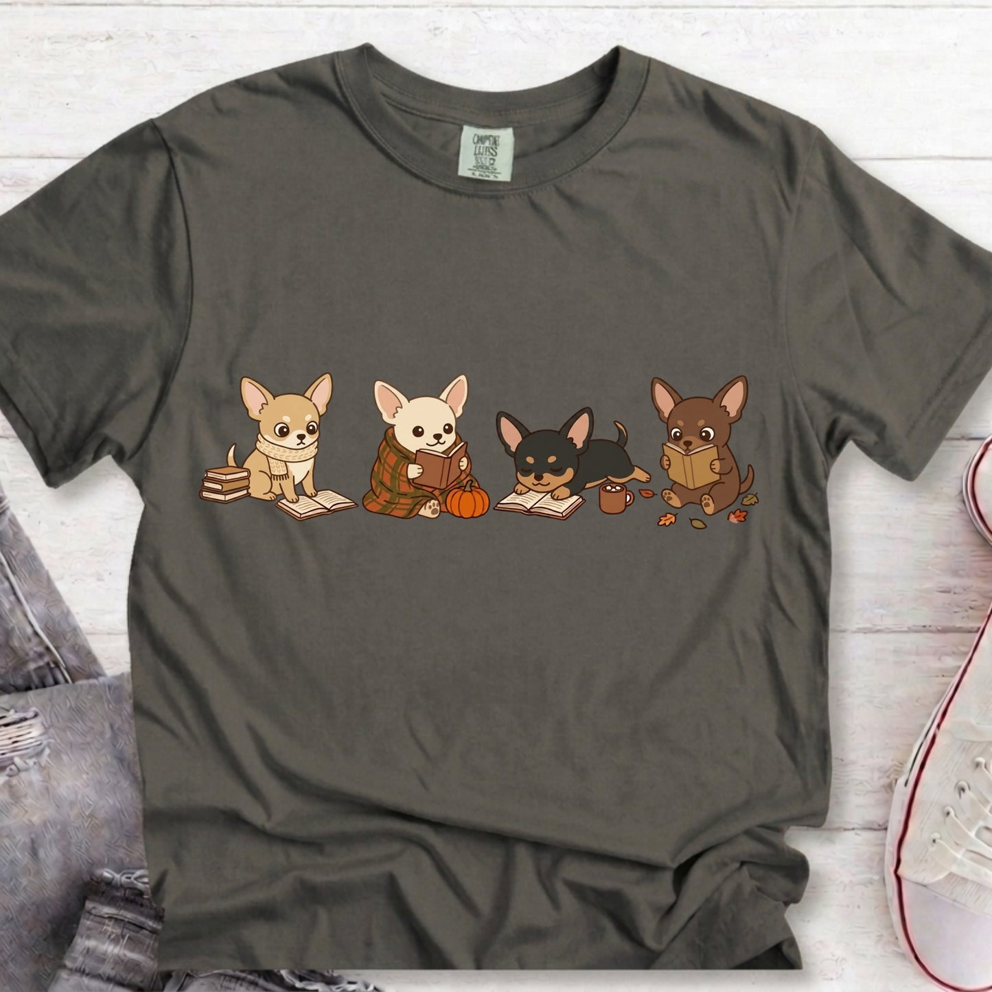 A dark gray t-shirt featuring a graphic of four cute cartoon Chihuahua dogs reading books, styled in a flat-lay on a white wood background with jeans and a sneaker partially visible.