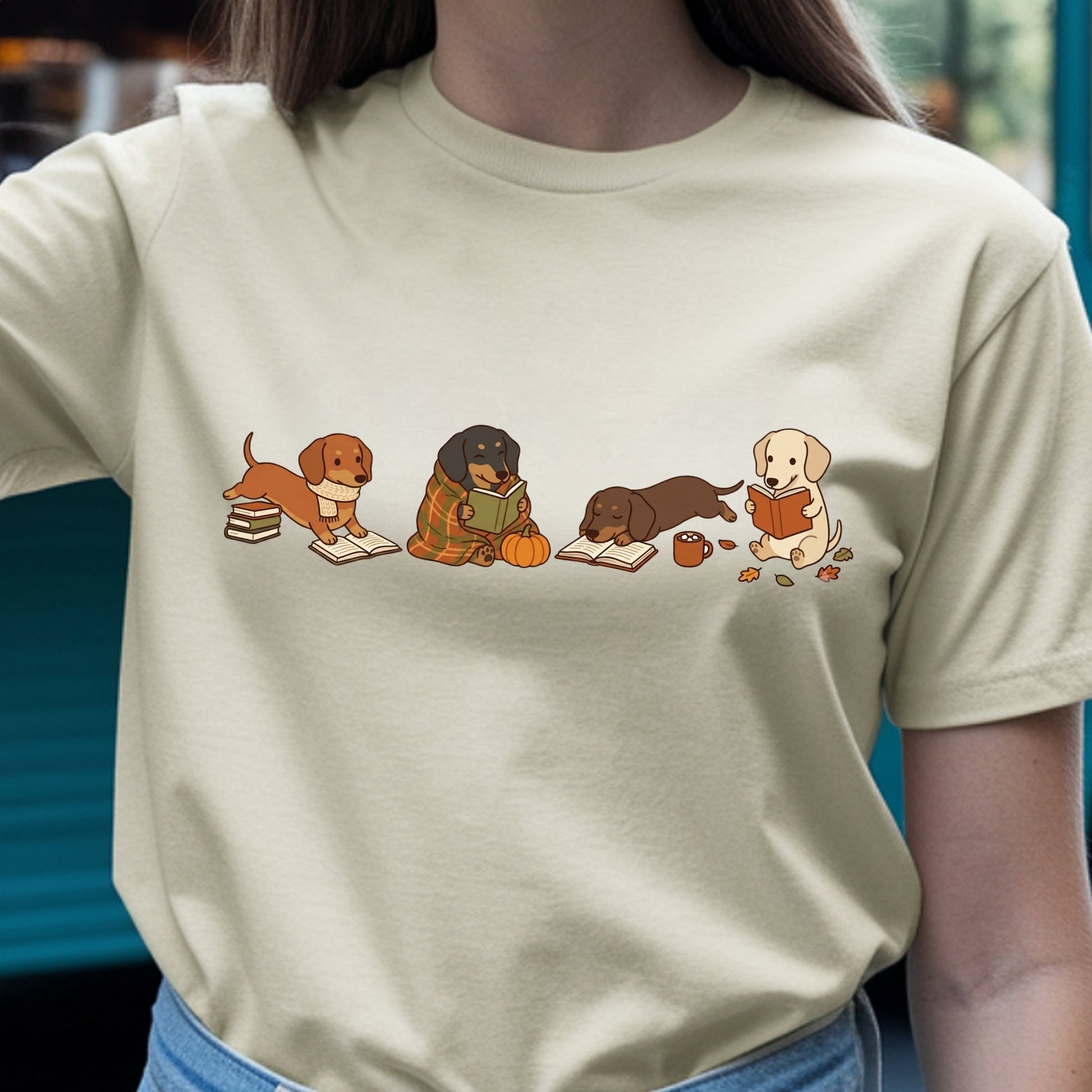 A person wearing a light beige crewneck t-shirt with a graphic print of four cute dachshund dogs reading books in an autumn setting.