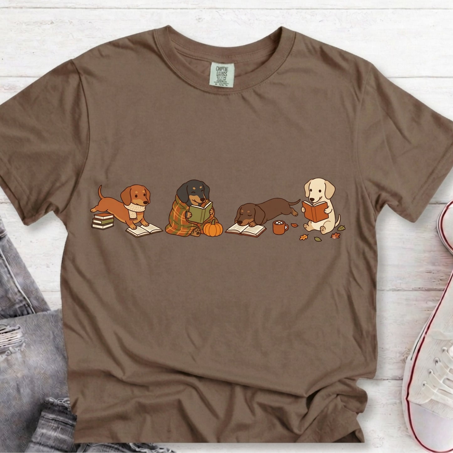 A brown short-sleeve crewneck t-shirt featuring a graphic of four dogs reading books, styled flat on a white wood background with jeans and a sneaker partially visible.