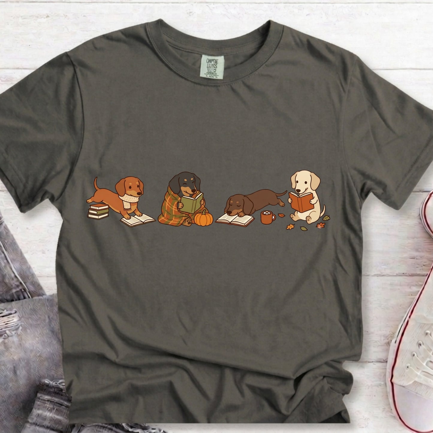A dark gray crewneck t-shirt featuring a graphic of four dachshunds sitting down and reading books, styled in a flat-lay on a white wood background with jeans and a sneaker partially visible.