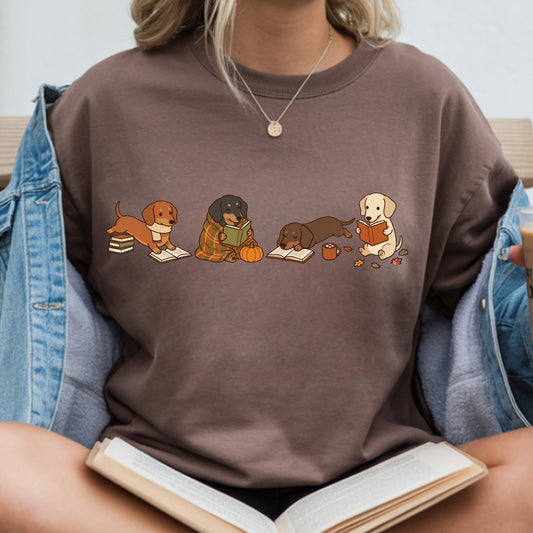 A person wearing a brown crewneck sweatshirt with a graphic of four dogs reading books. The person is also wearing an open denim jacket and holding an open book.