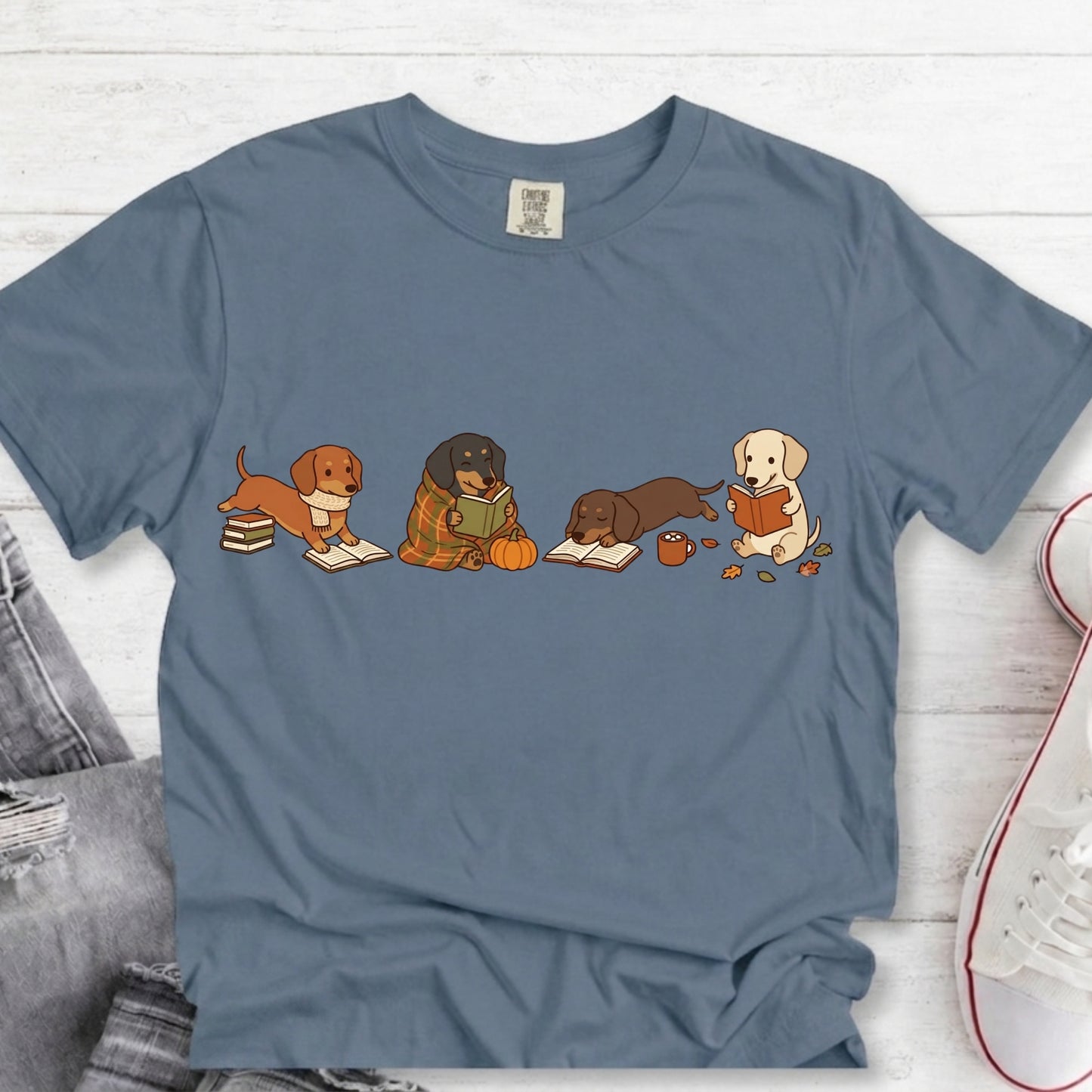 A slate blue crewneck t-shirt featuring a graphic of four dogs (three dachshunds and one yellow lab) reading books, styled in a flat lay on a white wood background with jeans and a sneaker partially visible.