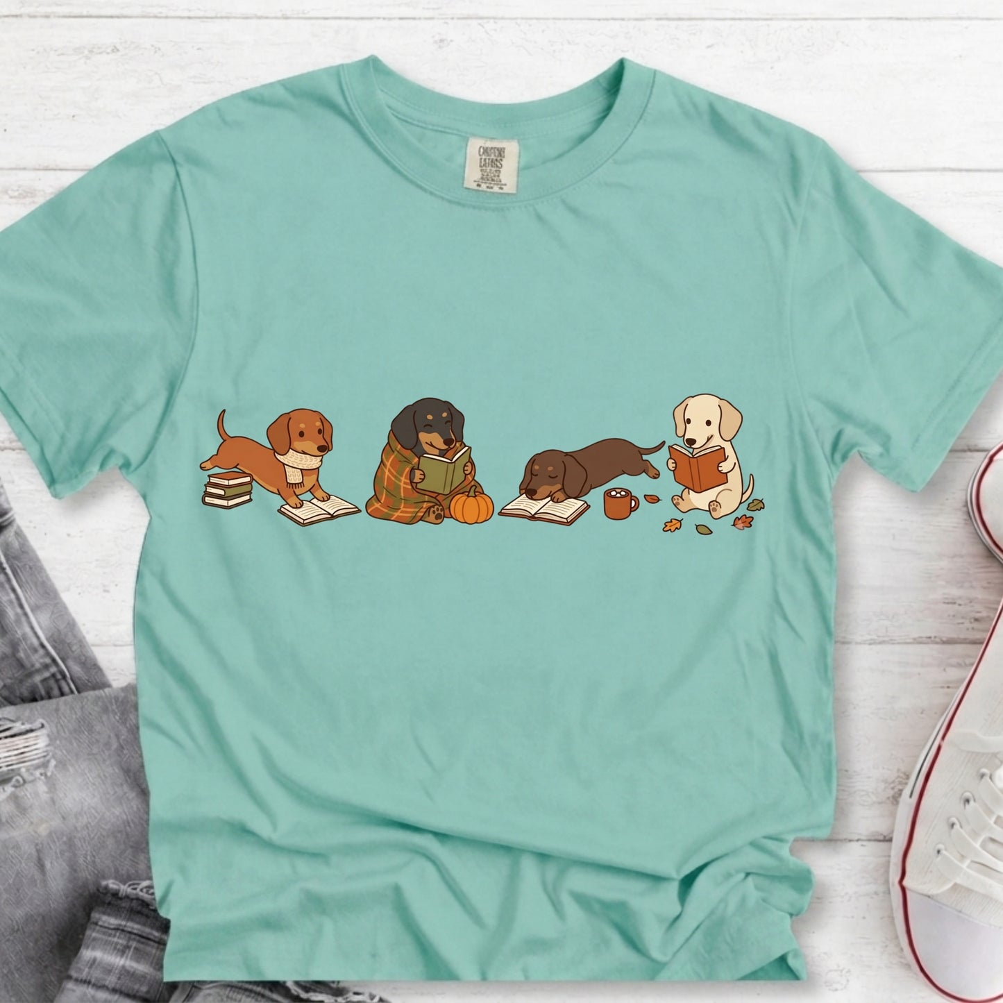 A mint green short-sleeve t-shirt with a graphic of four dogs reading books, laid flat on a white wood background with jeans and a sneaker partially visible.
