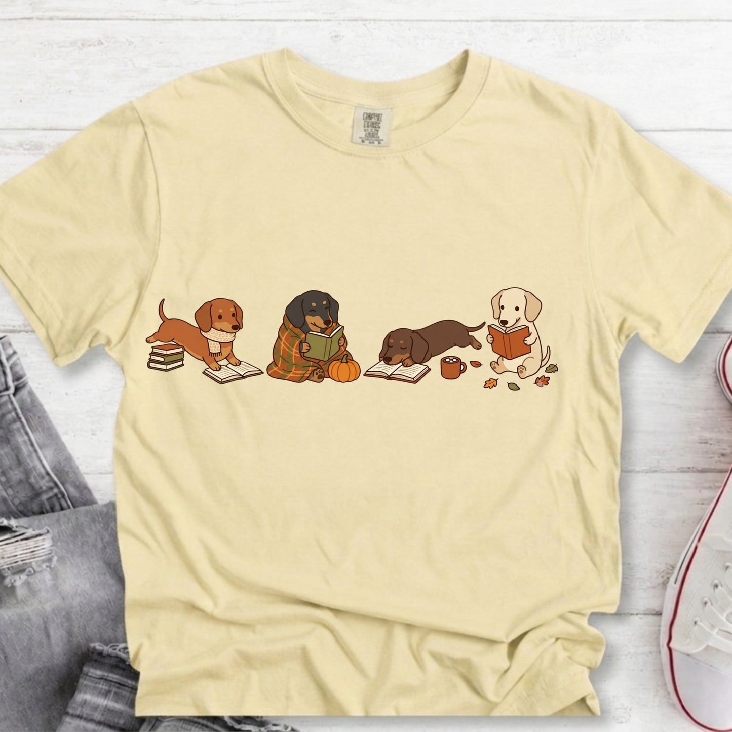 A pale yellow short-sleeve t-shirt with a graphic of four dachshunds reading books, styled in a flat-lay on a white wood background with jeans and a sneaker partially visible.