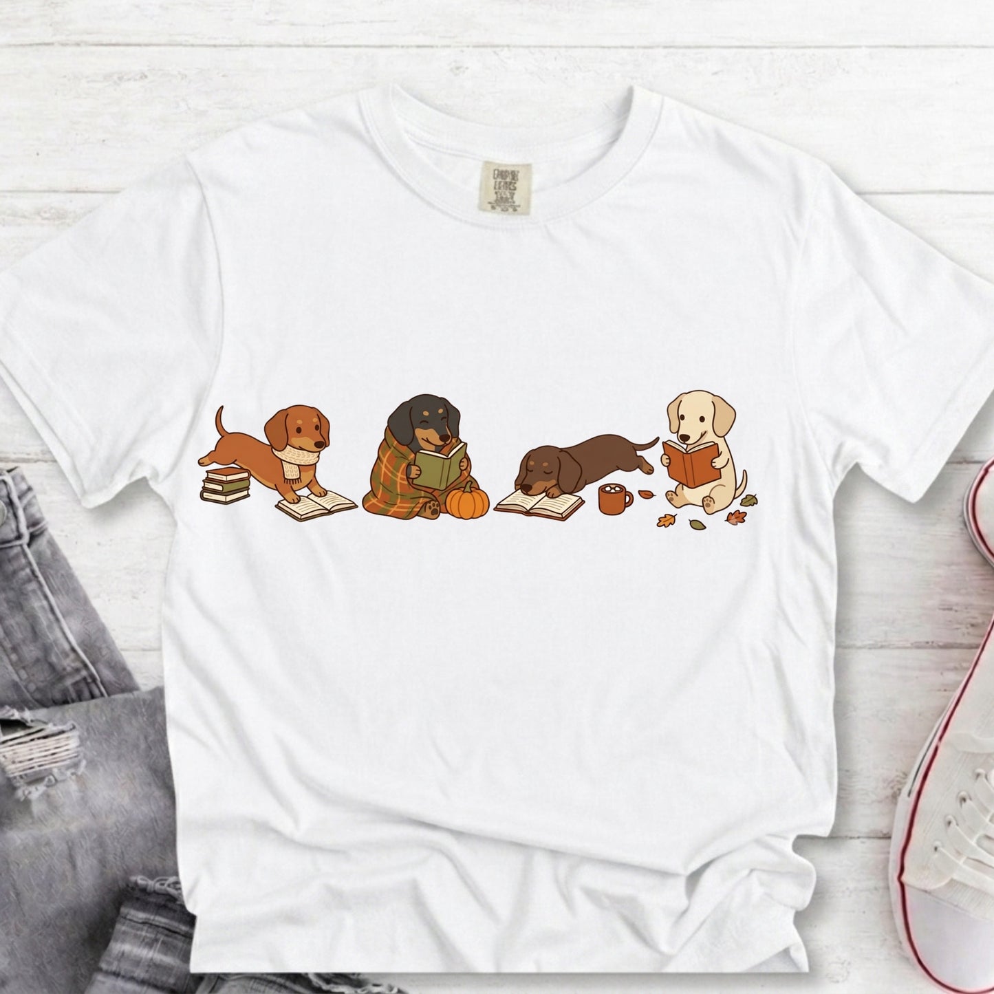 A white short-sleeve crewneck t-shirt featuring a graphic of four dachshunds reading books, styled in a flat-lay on a white wood background with jeans and a sneaker partially visible.