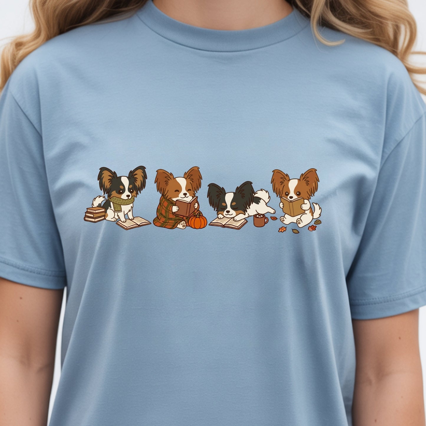 A light blue crewneck t-shirt featuring a graphic print of four cute Papillon dogs sitting in a row, each holding autumn-themed items like books, a pumpkin, and a cup. The t-shirt is shown on a model, cropped from the neck down.
