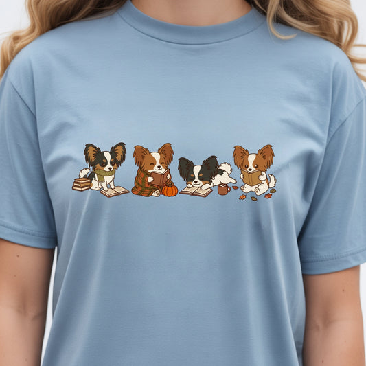 A light blue crewneck t-shirt featuring a graphic print of four cute Papillon dogs sitting in a row, each holding autumn-themed items like books, a pumpkin, and a cup. The t-shirt is shown on a model, cropped from the neck down.