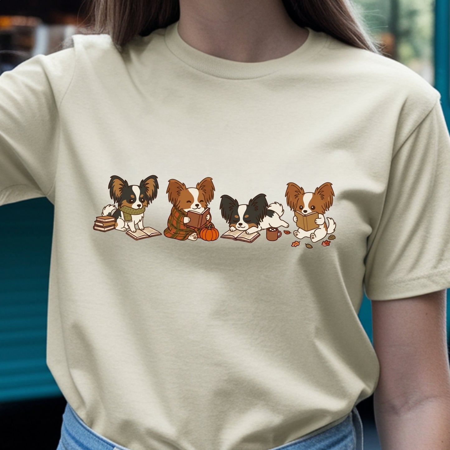 A person wearing a light beige crewneck t-shirt with a graphic print of five cute Papillon dogs reading books, sitting on a pumpkin, and playing with a small dog, paired with blue jeans.