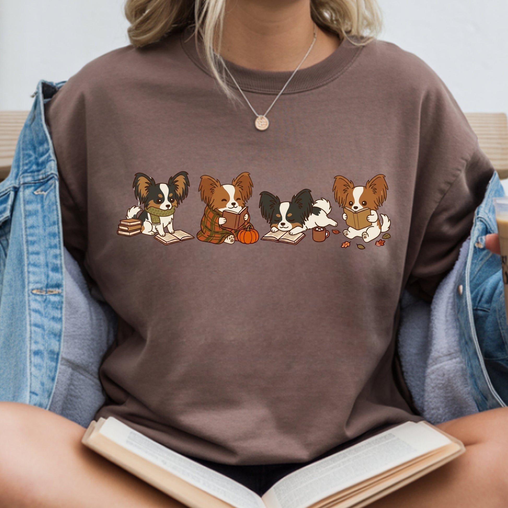A person wearing a brown crewneck sweatshirt with a graphic of four cartoon Papillon dogs sitting together reading books. The person is also wearing an open denim jacket and holding an open book.