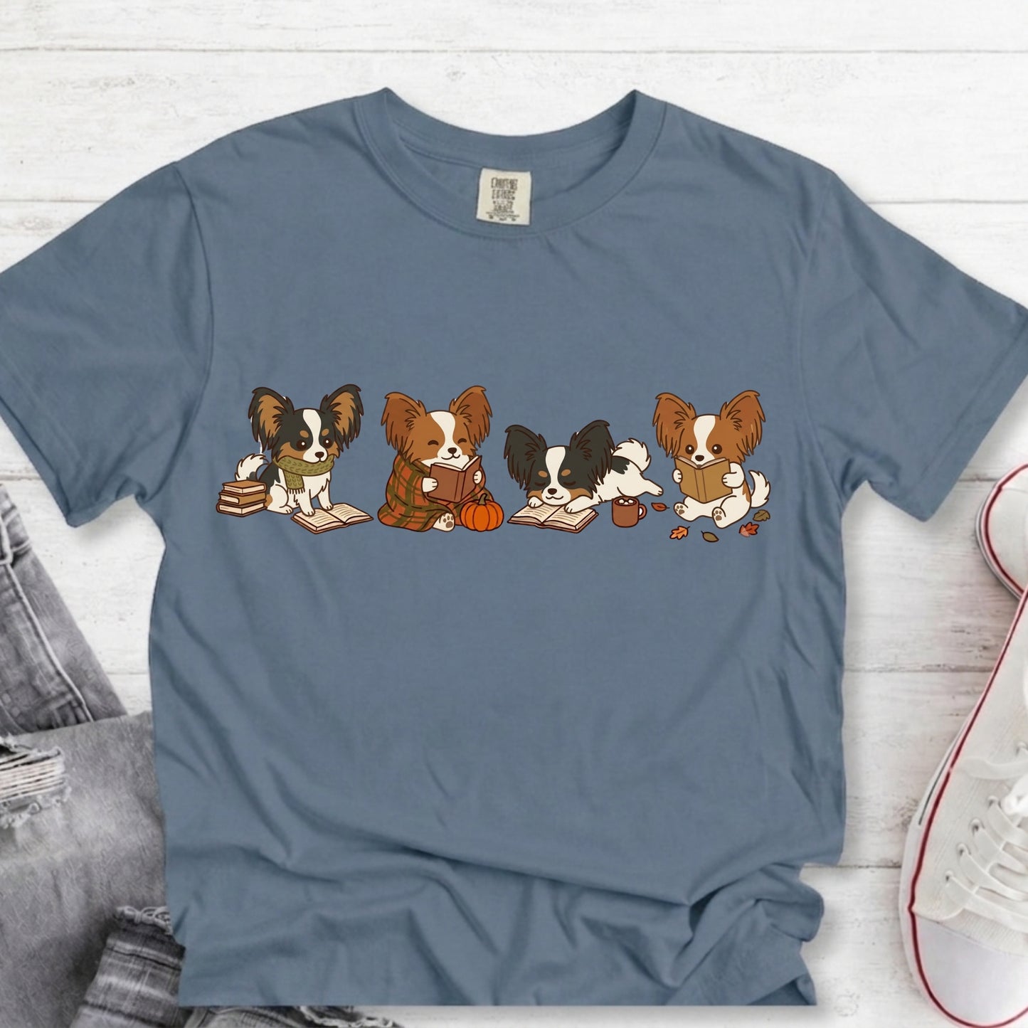 A slate blue crewneck t-shirt featuring a cartoon graphic of four Papillon dogs sitting on books and reading a pumpkin, laid flat on a white wood background with jeans and a sneaker partially visible.