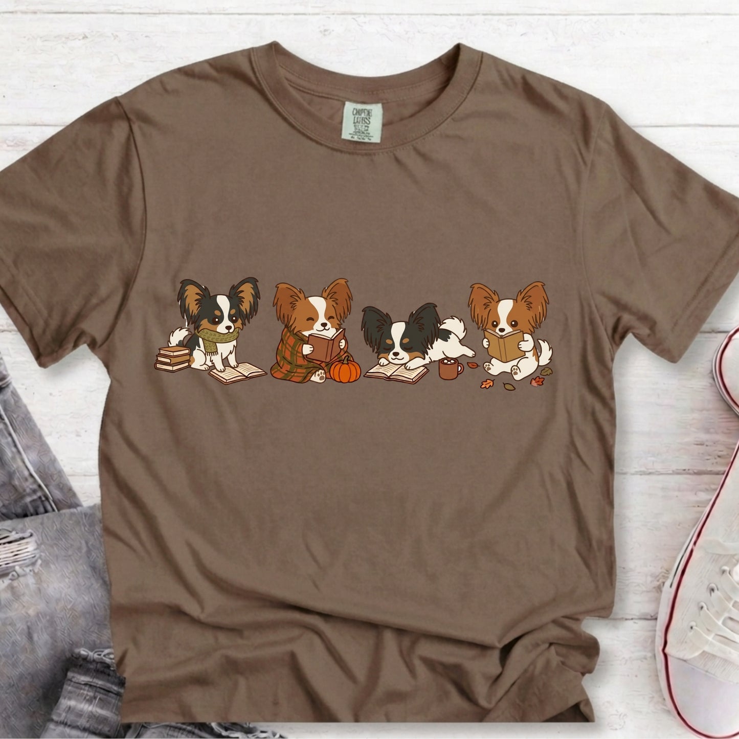 A brown short-sleeve t-shirt laid flat on a white wood background, featuring a graphic of four cartoon Papillon dogs sitting together reading books and holding a pumpkin. A pair of gray jeans and a white sneaker are partially visible as styling props.