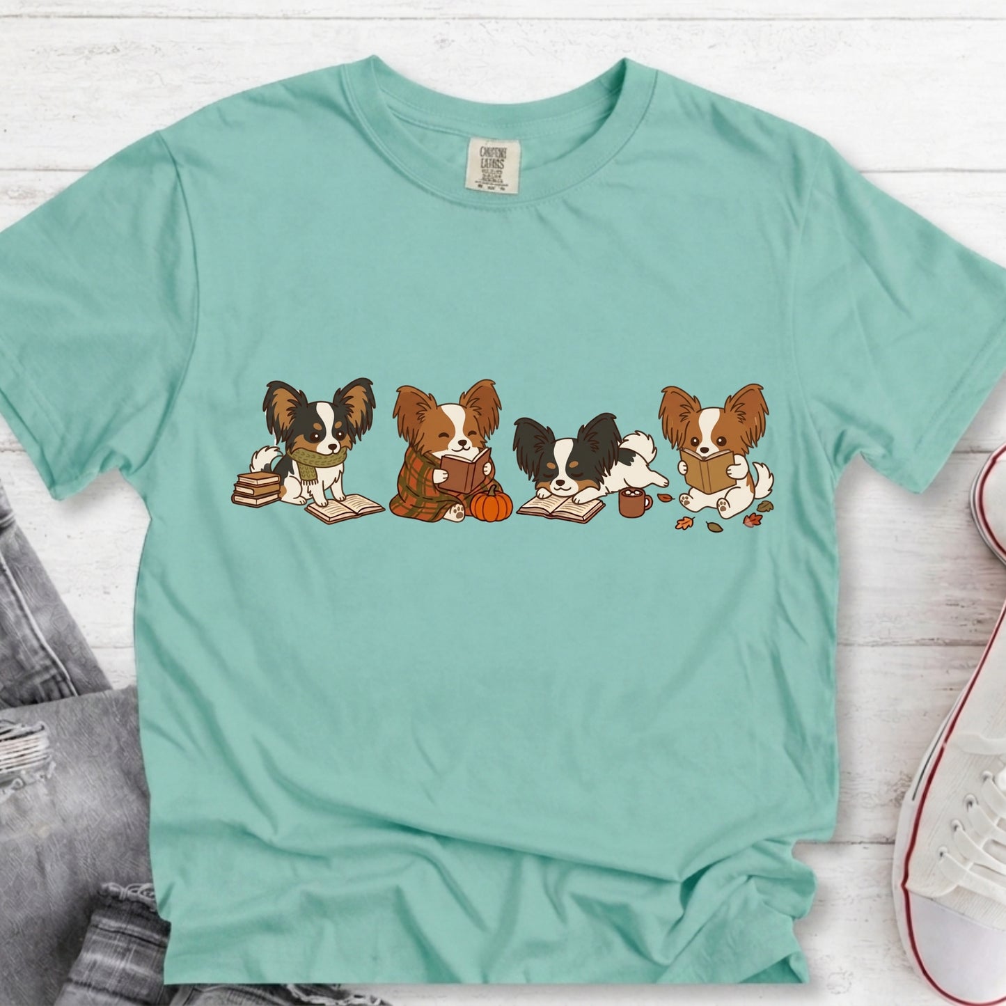 A mint green short-sleeve t-shirt with a graphic of four cartoon Papillon dogs sitting with books and an acorn, laid flat on a white wood background.