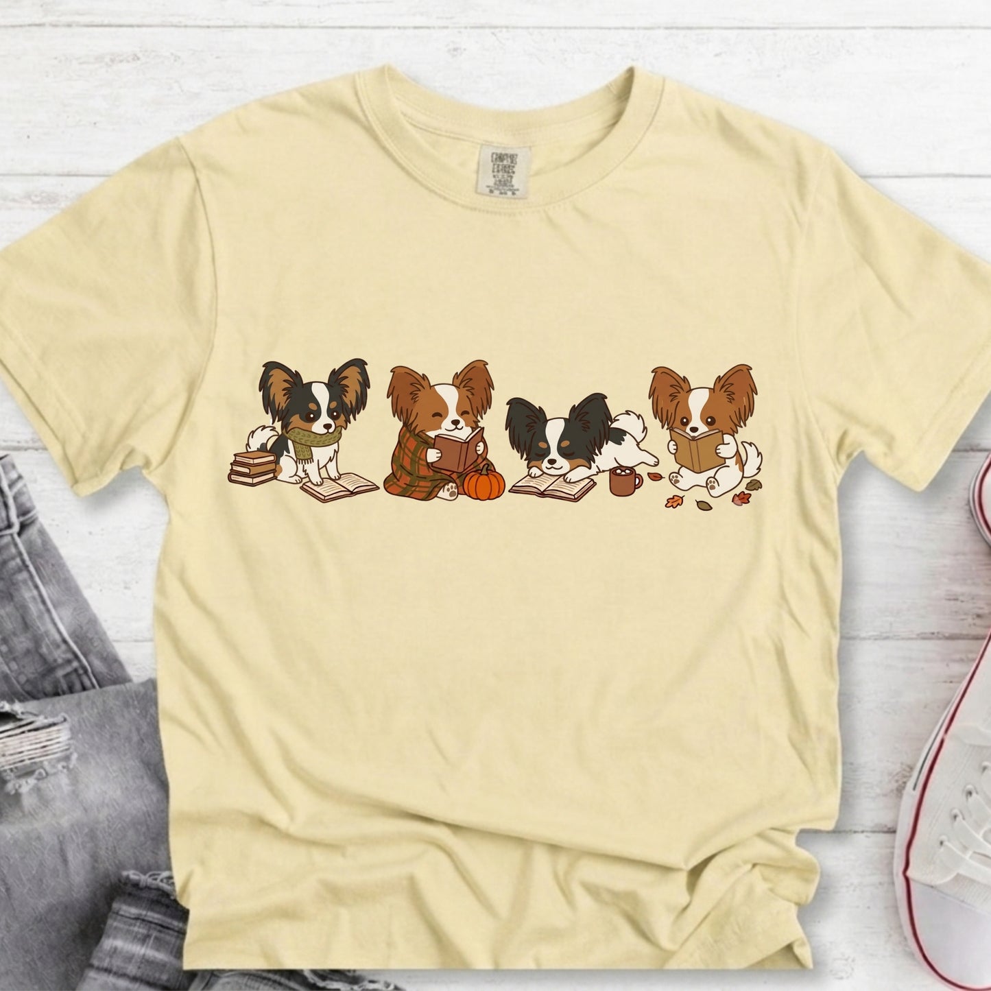 A pale yellow short-sleeve t-shirt laid flat on a white wood background, featuring a graphic print of four cartoon Papillon dogs sitting at books with a pumpkin and acorn.