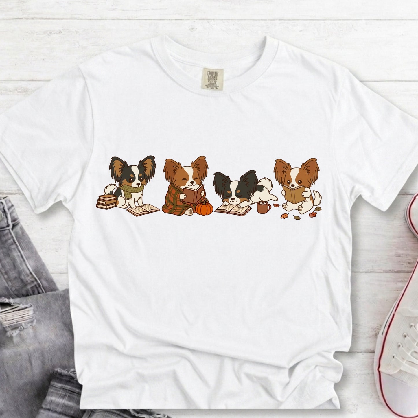 A white short-sleeve crewneck t-shirt laid flat on a light wood background, featuring a graphic of four cute cartoon Papillon dogs sitting on books and holding a pumpkin and acorn.