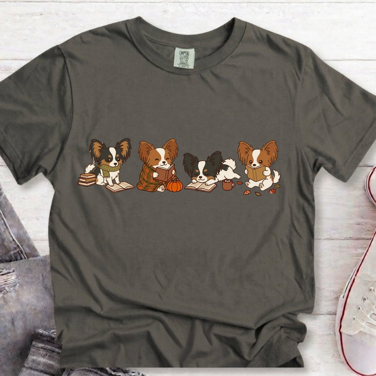 A dark gray t-shirt featuring a graphic of four cute cartoon Papillon dogs sitting and reading books, styled in a flat-lay on a white wood background with jeans and a sneaker partially visible.