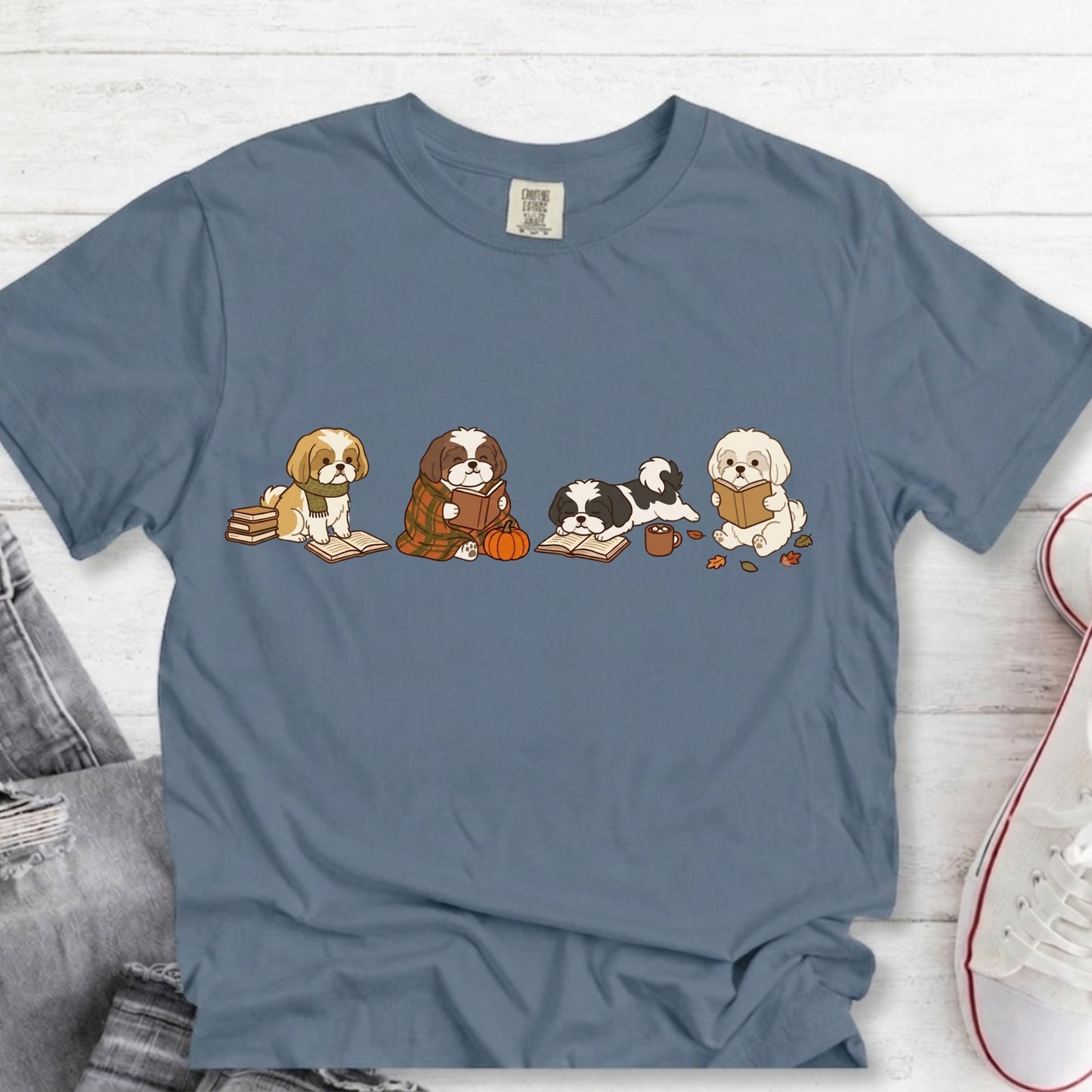 A slate blue crewneck t-shirt featuring a graphic of five cute cartoon dogs sitting on books and a pumpkin, laid flat on a white wood background with jeans and a sneaker partially visible.