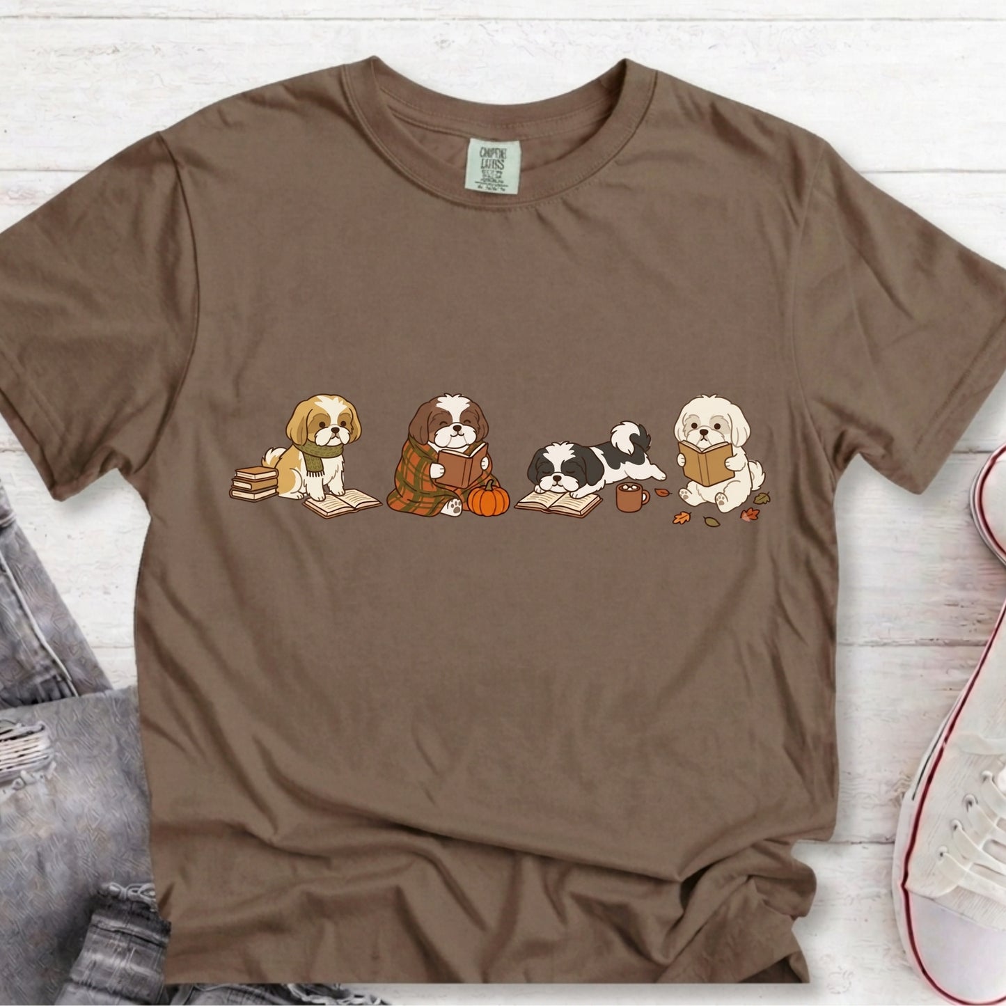 A brown short-sleeve crewneck t-shirt featuring a graphic print of four cute cartoon dogs sitting with books and a pumpkin. The shirt is laid flat on a white wood background with a denim jacket and a sneaker partially visible as styling props.