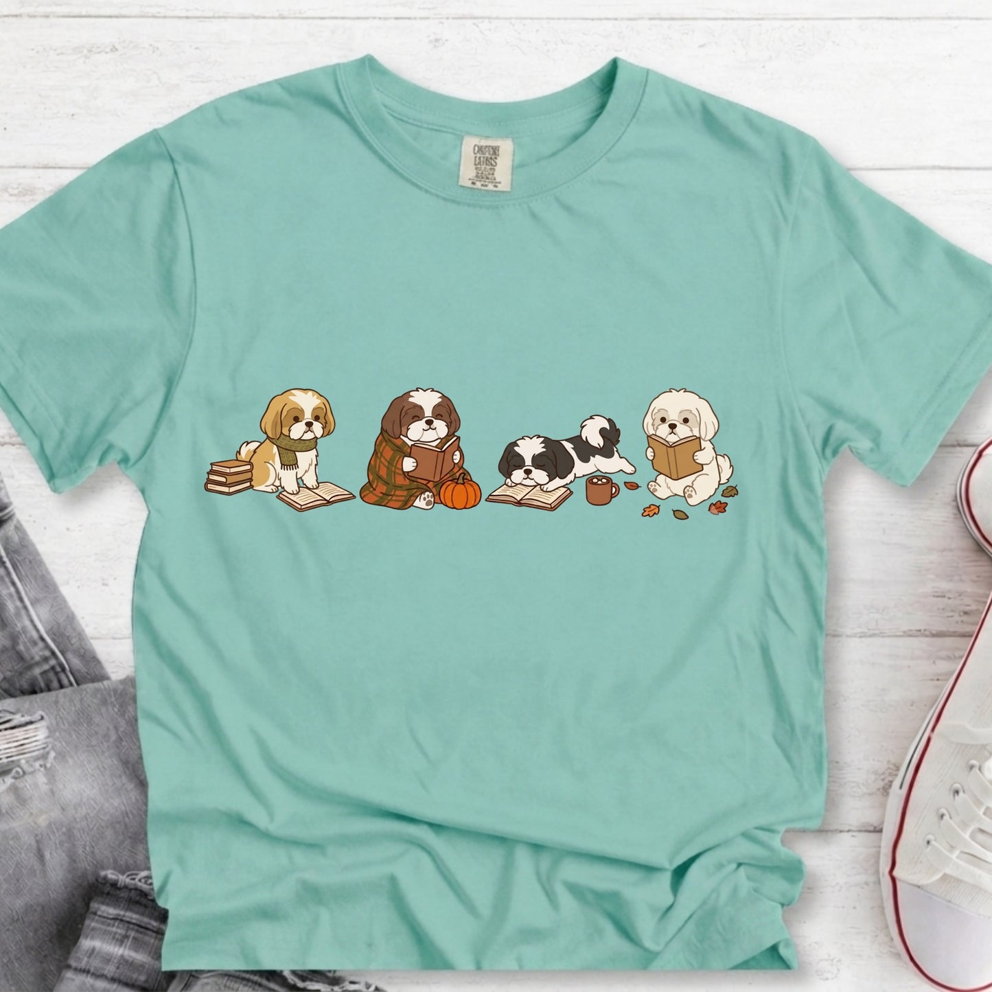 A mint green short-sleeve t-shirt with a graphic of five cute cartoon dogs reading books, laid flat on a white wood background with jeans and a sneaker partially visible.