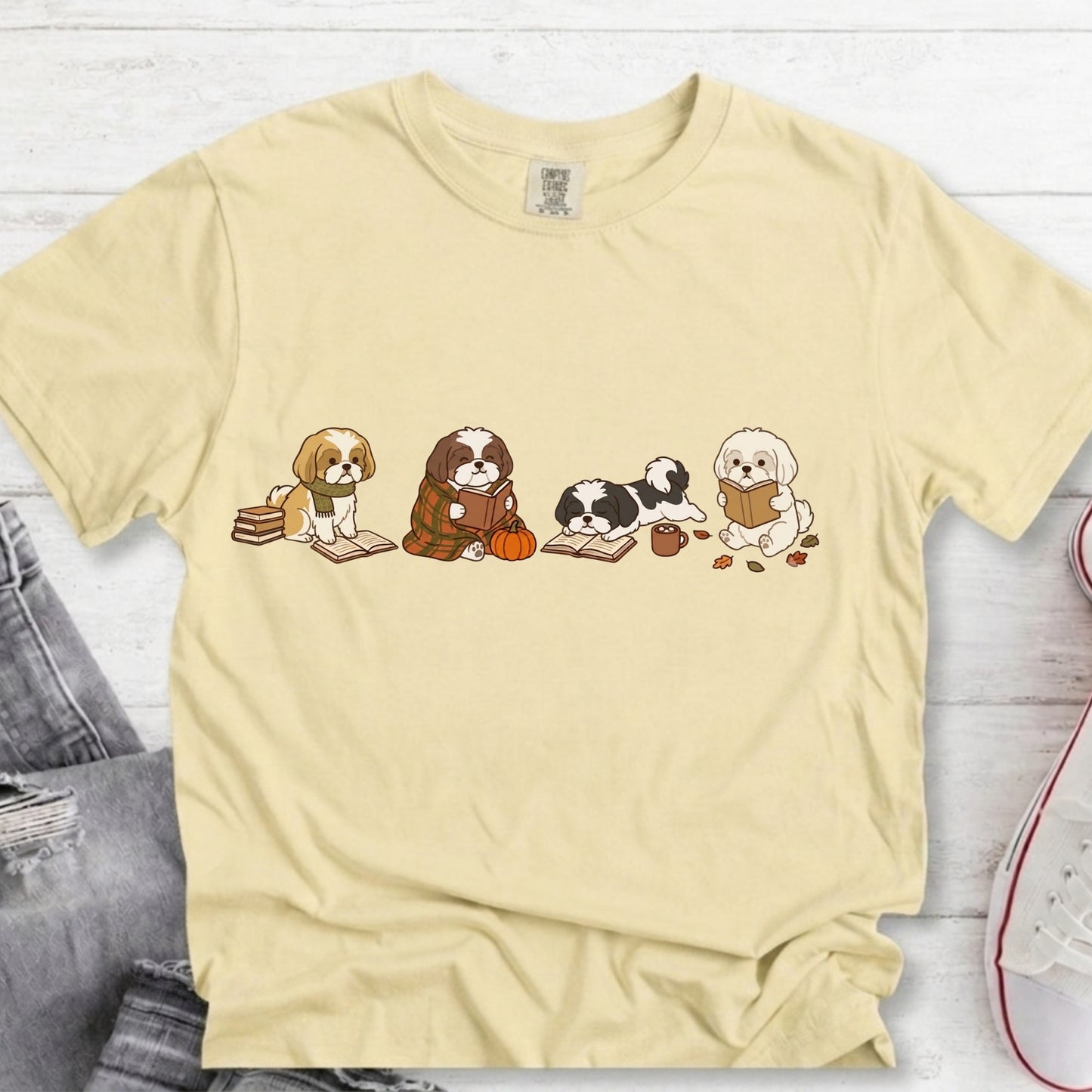 A pale yellow short-sleeve t-shirt with a graphic of four cute cartoon dogs reading books, styled in a flat-lay on a white wood background with jeans and a sneaker partially visible.