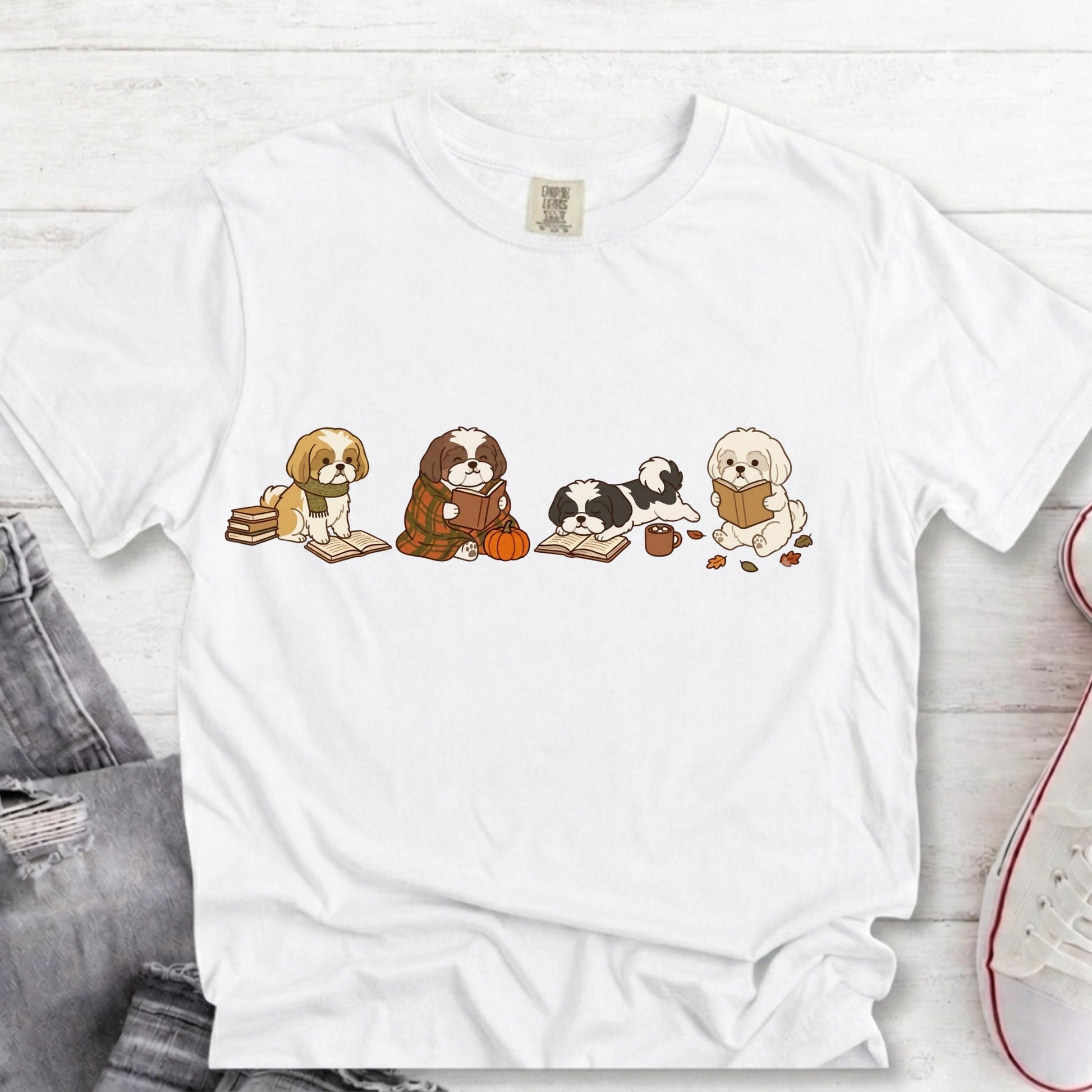 A white short-sleeve crewneck t-shirt featuring a graphic print of four cute dog puppies reading books, styled in a flat-lay arrangement on a white wood background with jeans and a sneaker partially visible.