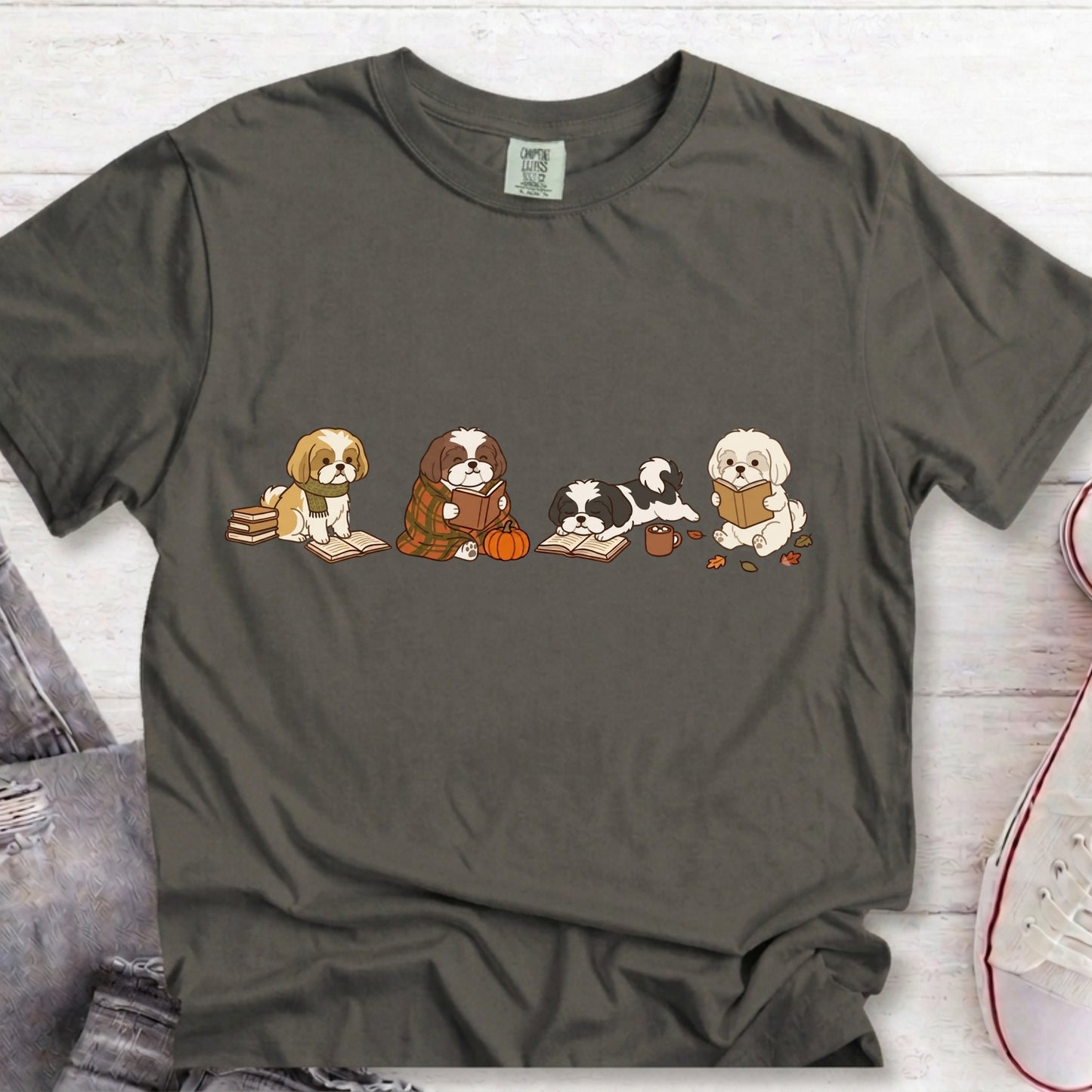A dark gray t-shirt with a graphic of four cute cartoon dogs reading books, styled in a flat-lay on a white wood background with jeans and a sneaker partially visible.