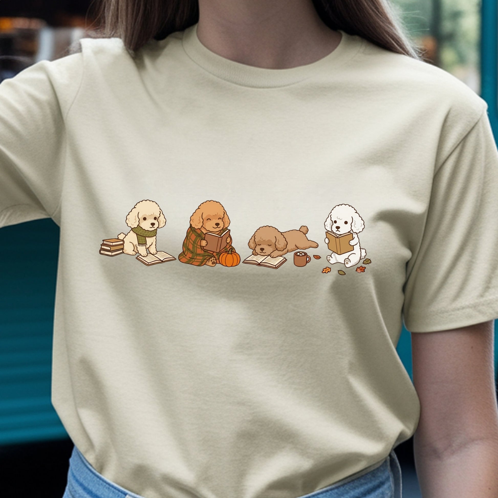 A person wearing a light beige crewneck t-shirt with a graphic of four cute cartoon dogs reading books on a fall-themed background.
