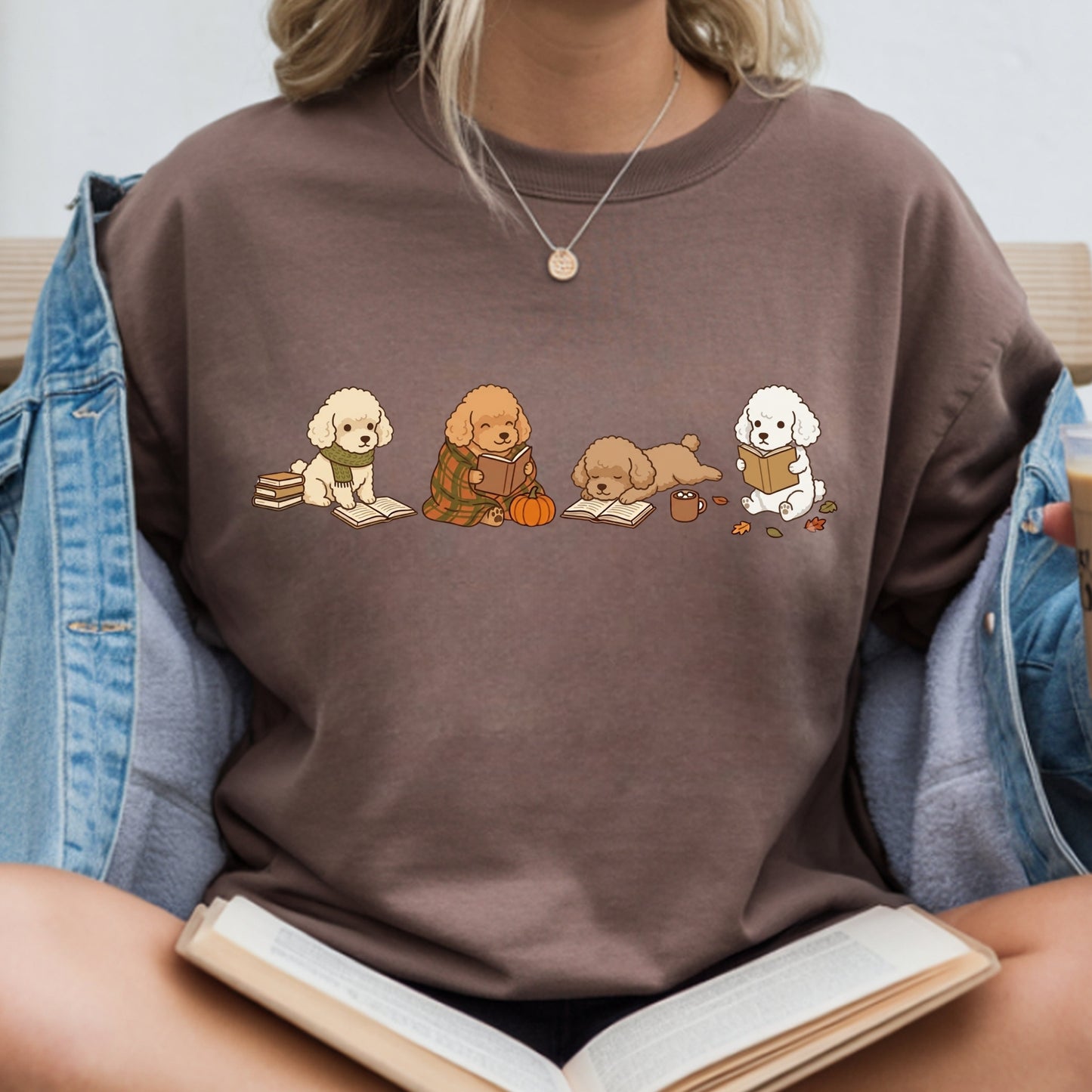 A person wearing a brown crewneck sweatshirt with a cute graphic of four dogs reading books, styled with an open denim jacket and holding an open book.