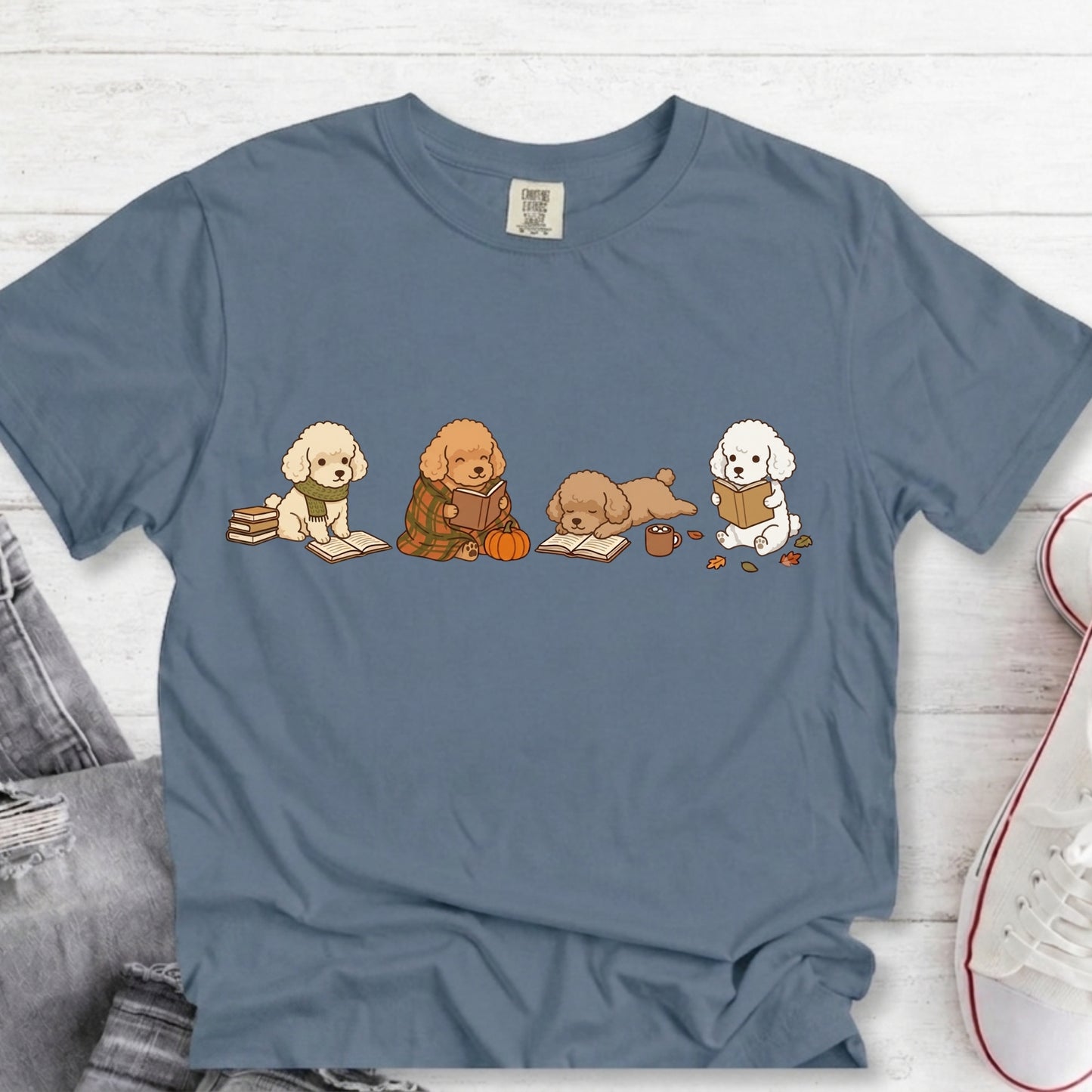 A slate blue crewneck t-shirt featuring a graphic of four cute cartoon dogs reading books, styled in a flat-lay on a white wood background with jeans and a sneaker partially visible.