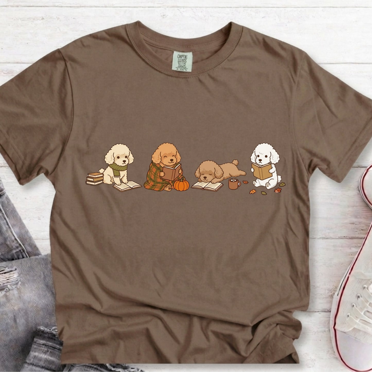 A brown short-sleeve crewneck t-shirt featuring a graphic print of four cute dogs reading books, styled in a flat-lay on a white wood background with jeans and a sneaker partially visible.