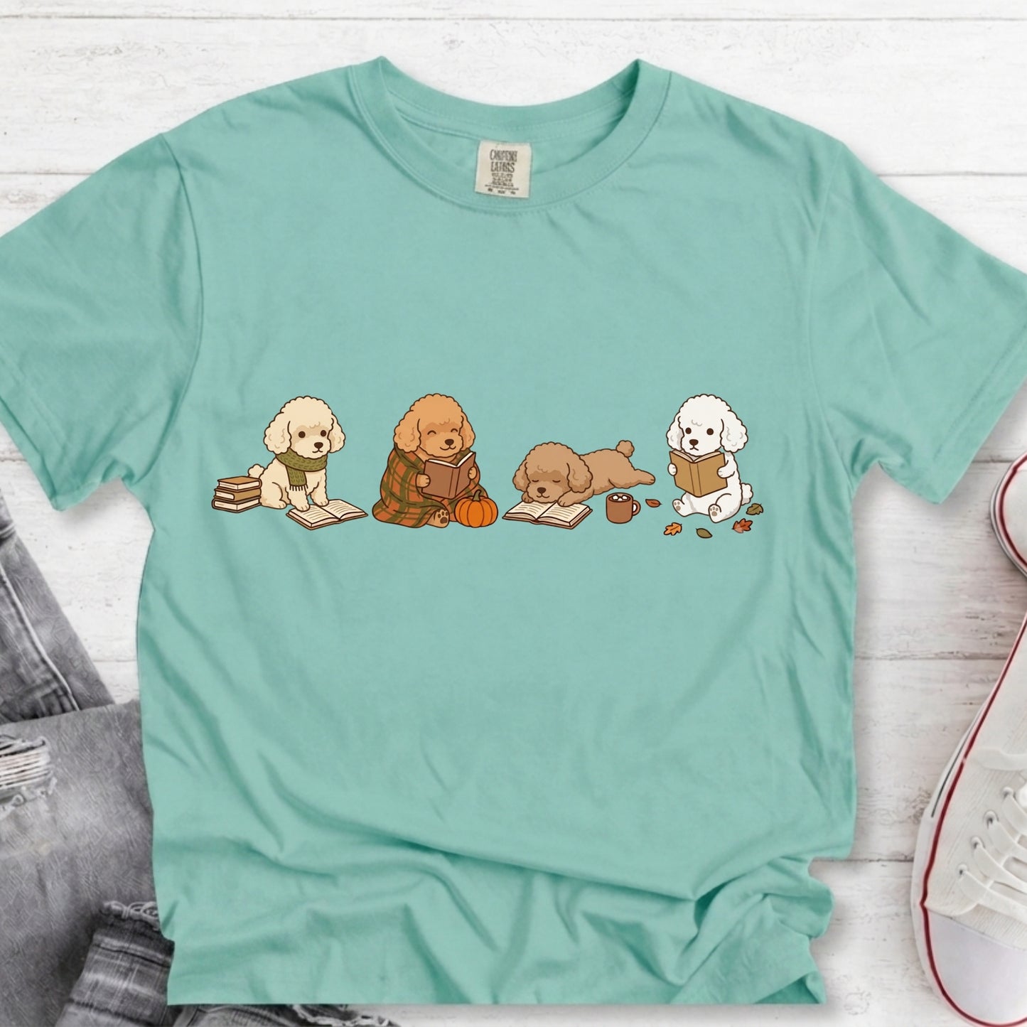 A mint green short-sleeve t-shirt laid flat on a white wood background, featuring a graphic of four cute cartoon dogs sitting and reading. The t-shirt is styled with a pair of gray jeans and a white sneaker in the corners.