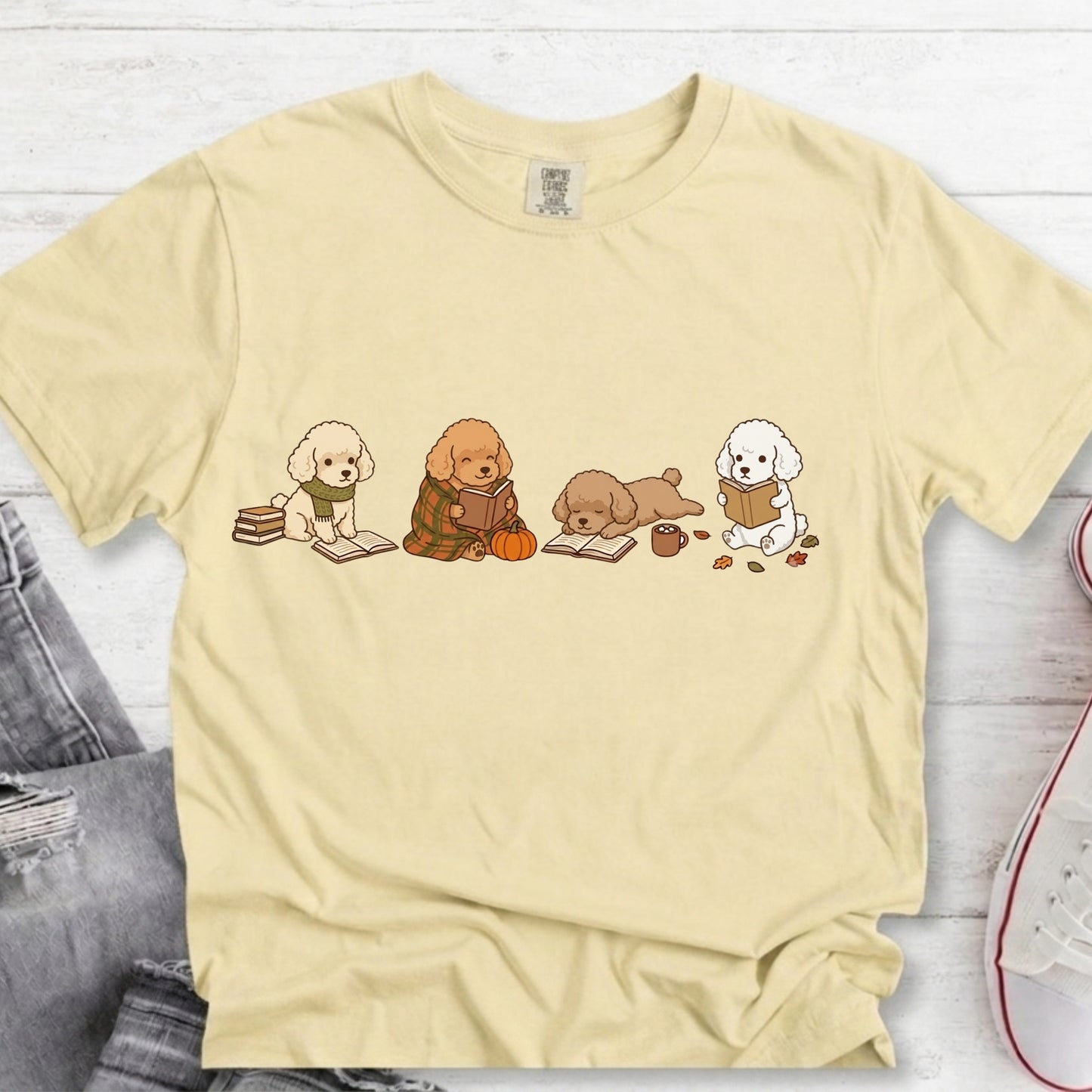 A pale yellow short-sleeve t-shirt with a graphic of three cute cartoon poodles reading books, styled in a flat-lay on a white wood background with jeans and a sneaker partially visible.