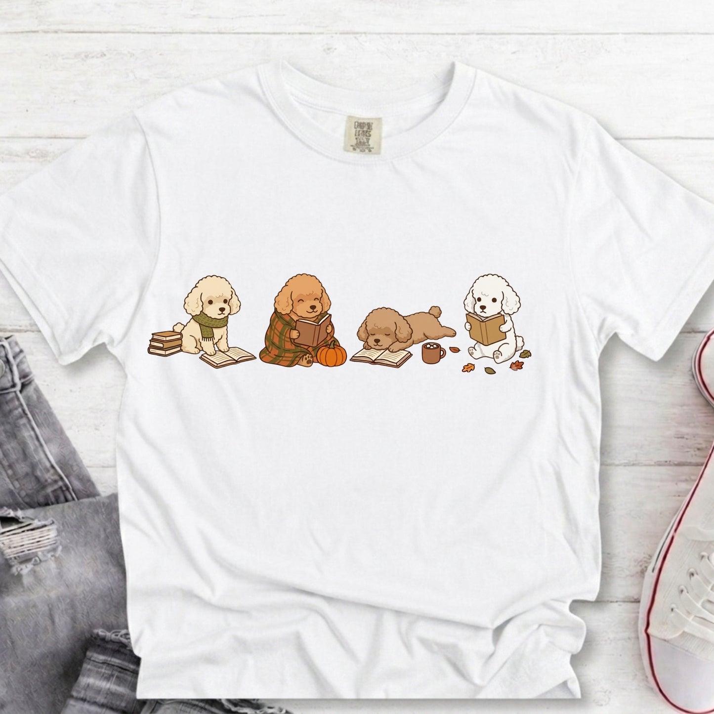 A white short-sleeve crewneck t-shirt featuring a graphic print of four cute cartoon dog puppies reading books, styled in a flat-lay on a white wood background with jeans and a sneaker partially visible.