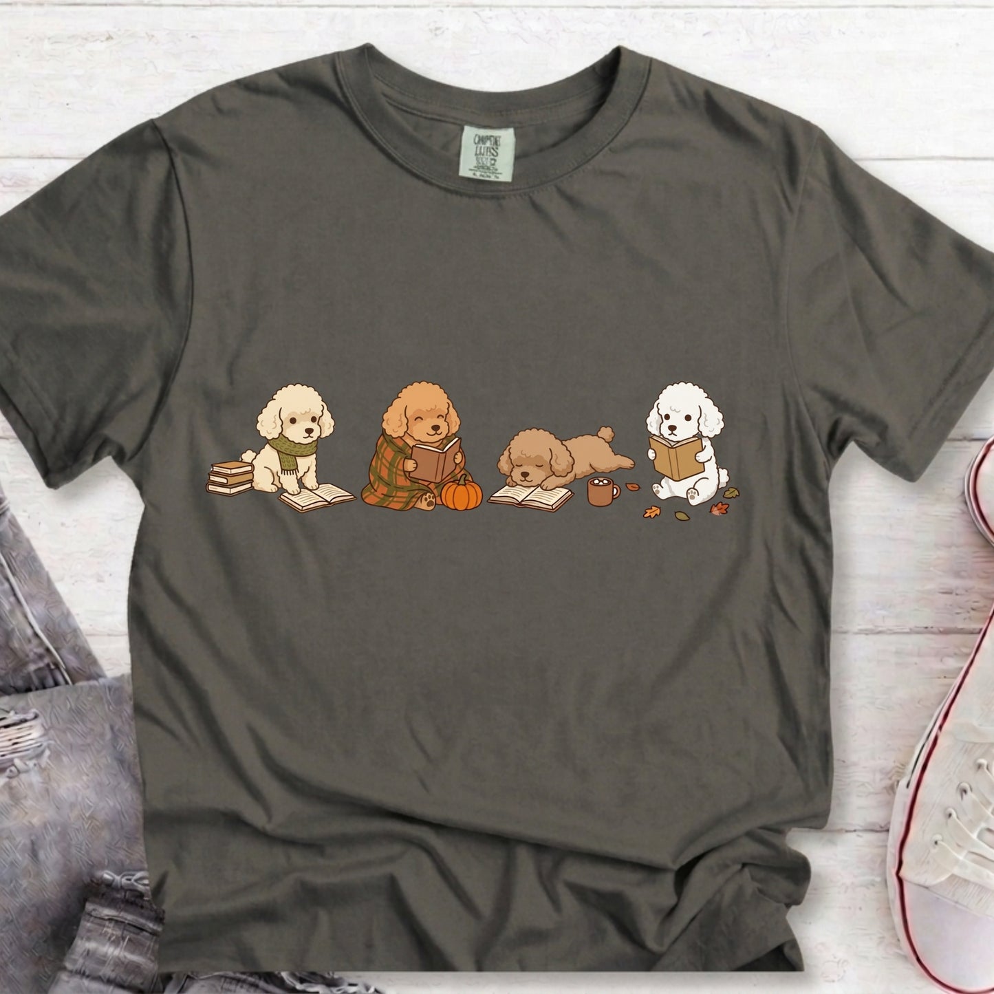 A dark gray t-shirt with a graphic of four cute cartoon dogs reading books, styled in a flat-lay on a white wood background with jeans and a sneaker partially visible.