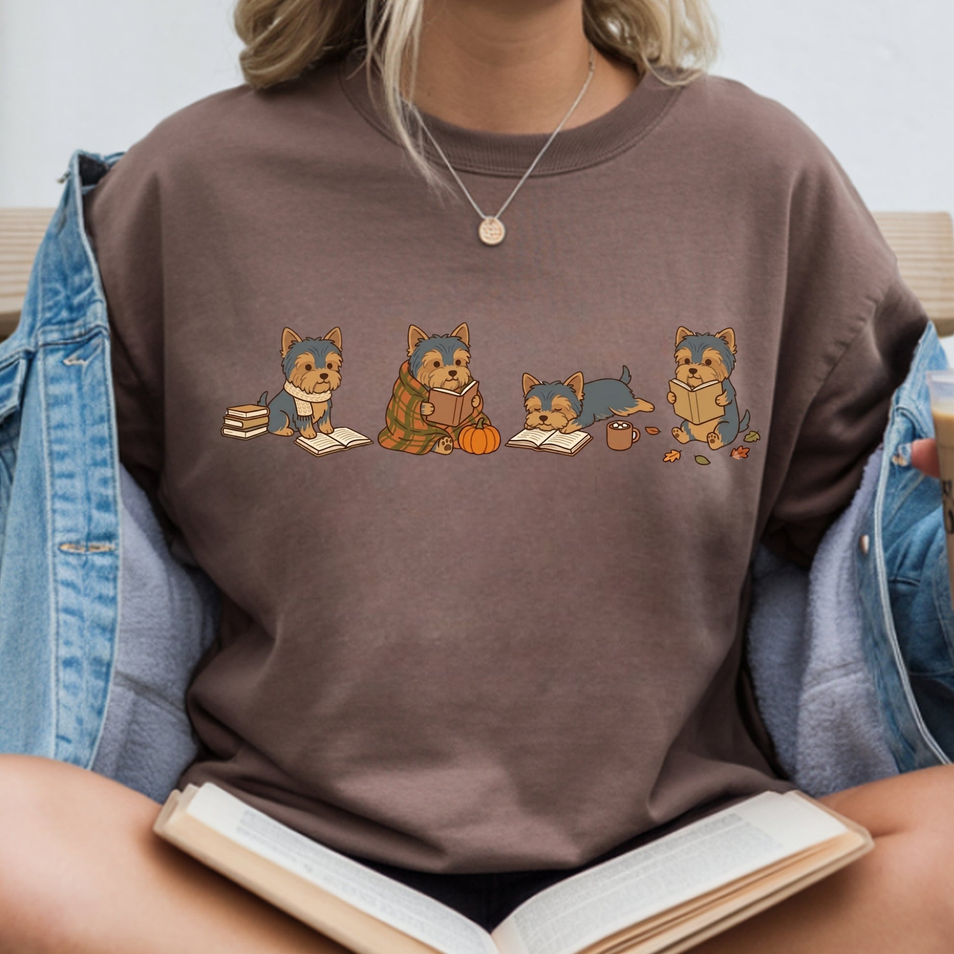 A person wearing a brown crewneck sweatshirt with a graphic of four small dogs reading books, styled with an open denim jacket and holding an open book.