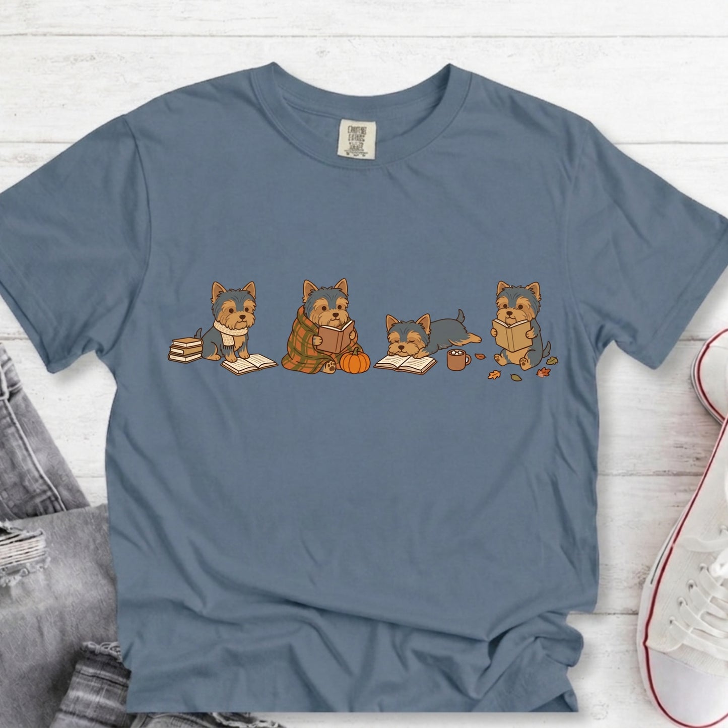 A slate blue crewneck t-shirt featuring a graphic of four cute German Shepherd dogs sitting and reading, styled in a flat-lay on a white wood background with jeans and a sneaker partially visible.