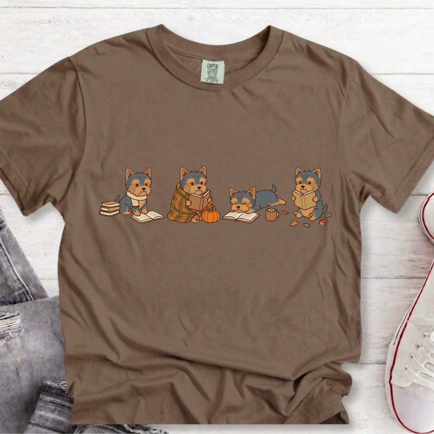 A brown short-sleeve crewneck t-shirt featuring a graphic of four cute cartoon dogs dressed in autumn clothing sitting with books and a pumpkin. The shirt is laid flat on a white wood background with jeans and a sneaker partially visible as styling props.