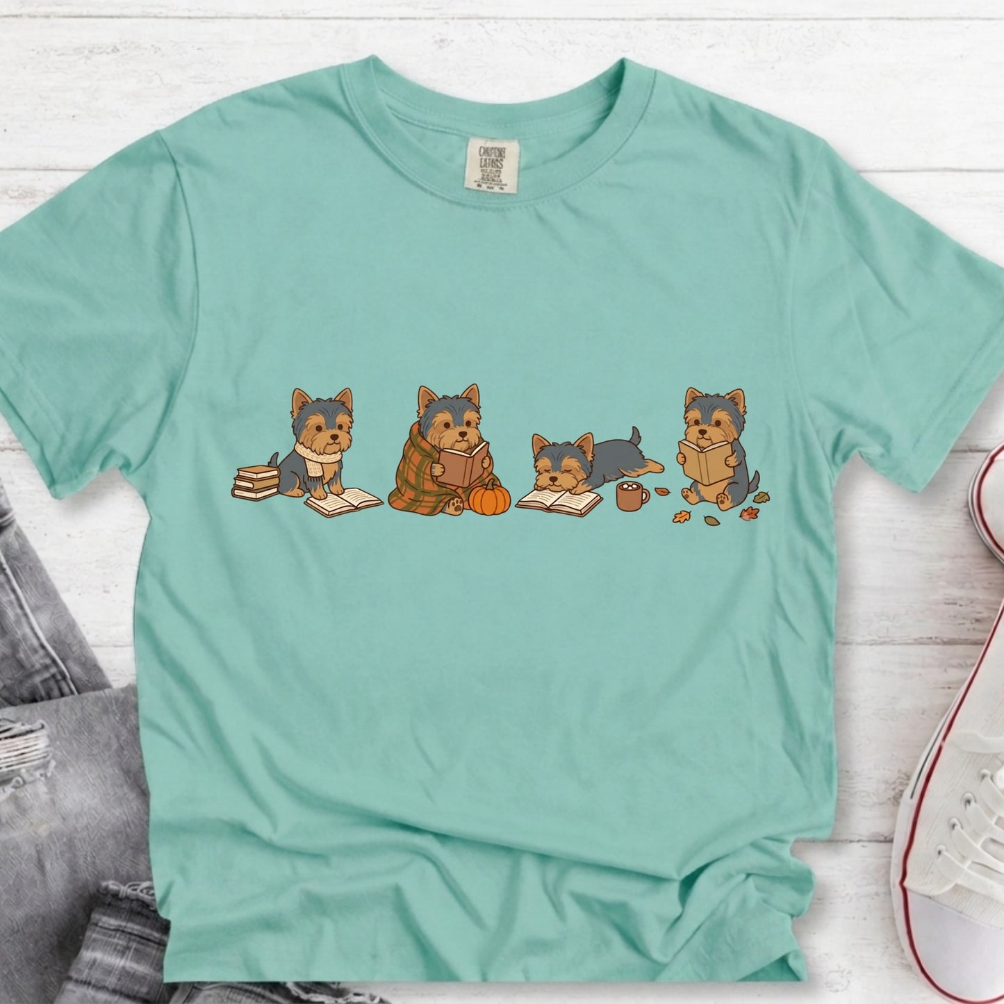 A mint green short-sleeve t-shirt with a graphic of four cartoon Yorkshire Terriers sitting with books and a pumpkin, laid flat on a white wood background with jeans and a sneaker partially visible.