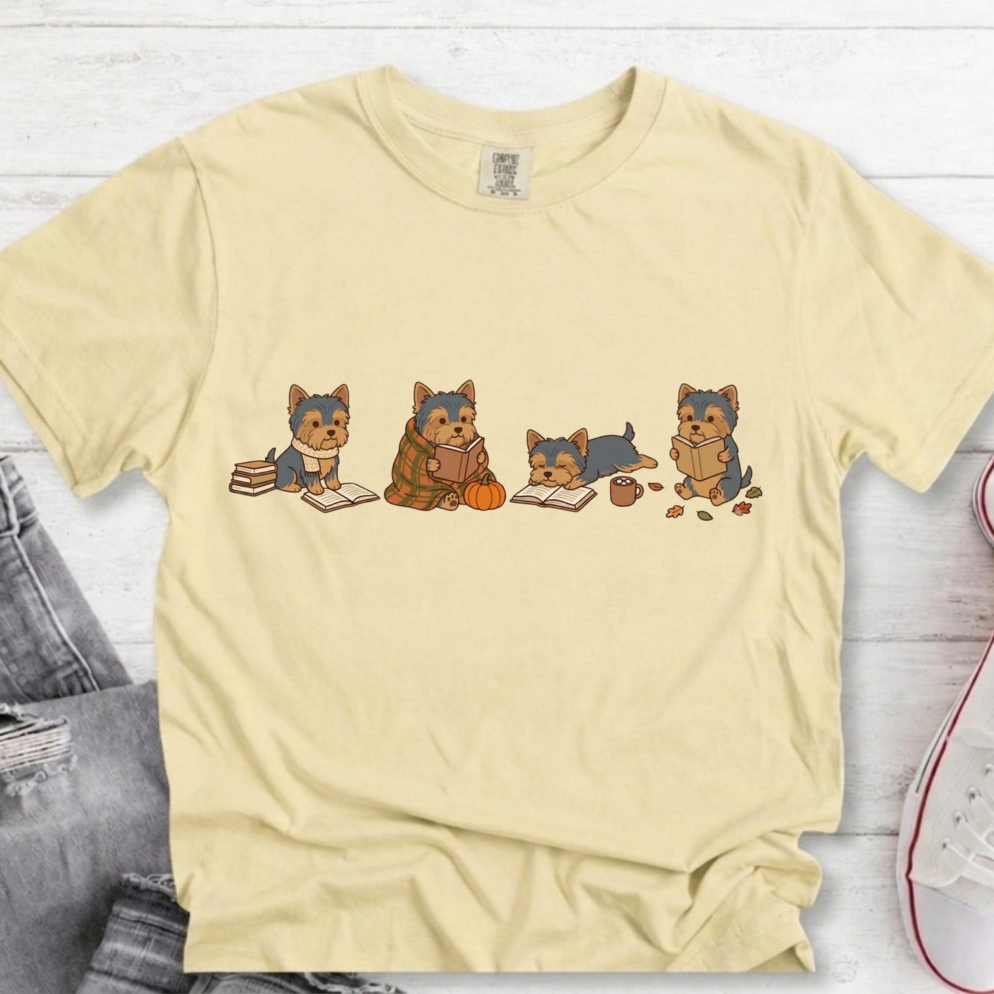 A pale yellow short-sleeve t-shirt laid flat on a white wood background, featuring a graphic of four cartoon Yorkshire Terrier dogs sitting with books and treats. Gray jeans and a white sneaker are partially visible at the edges as styling props.