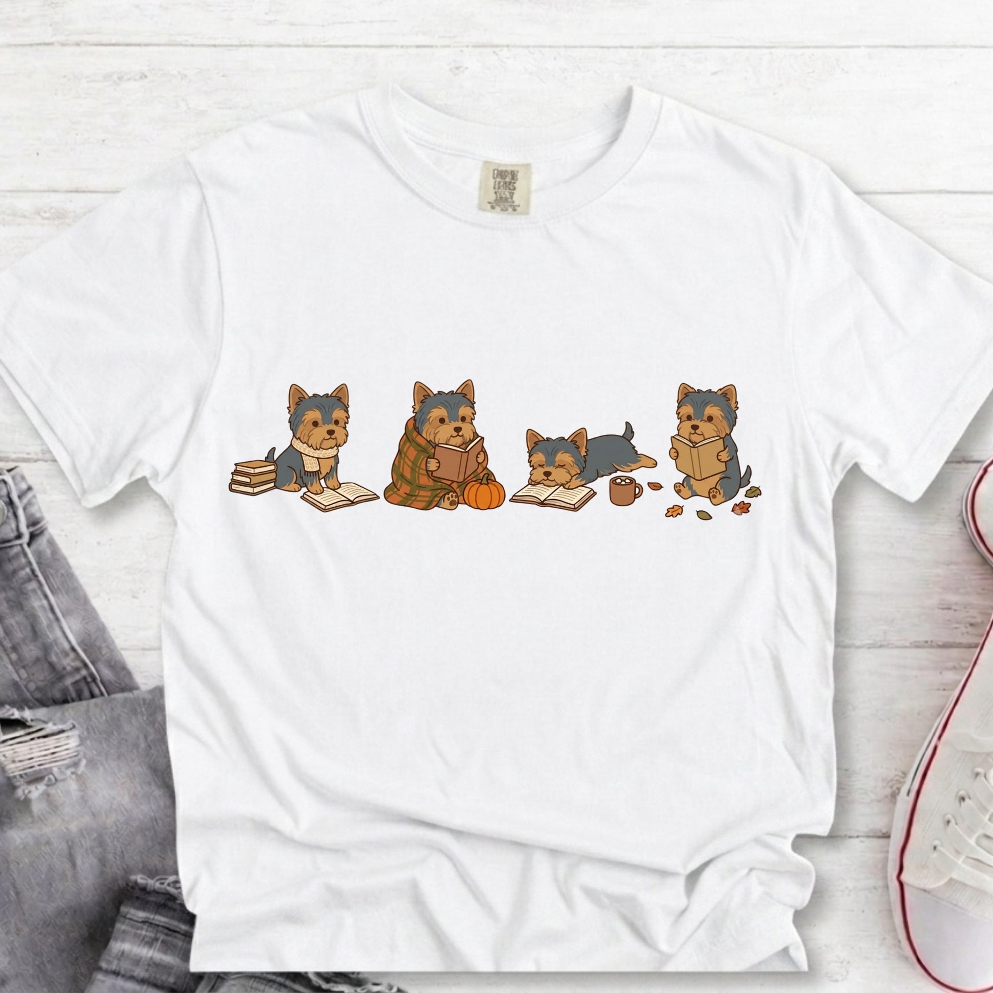 A white short-sleeve crewneck t-shirt featuring a graphic print of four cartoon Yorkshire Terrier dogs sitting with books and a pumpkin. The shirt is displayed in a flat-lay arrangement on a white wood background, partially styled with gray jeans and a white sneaker.