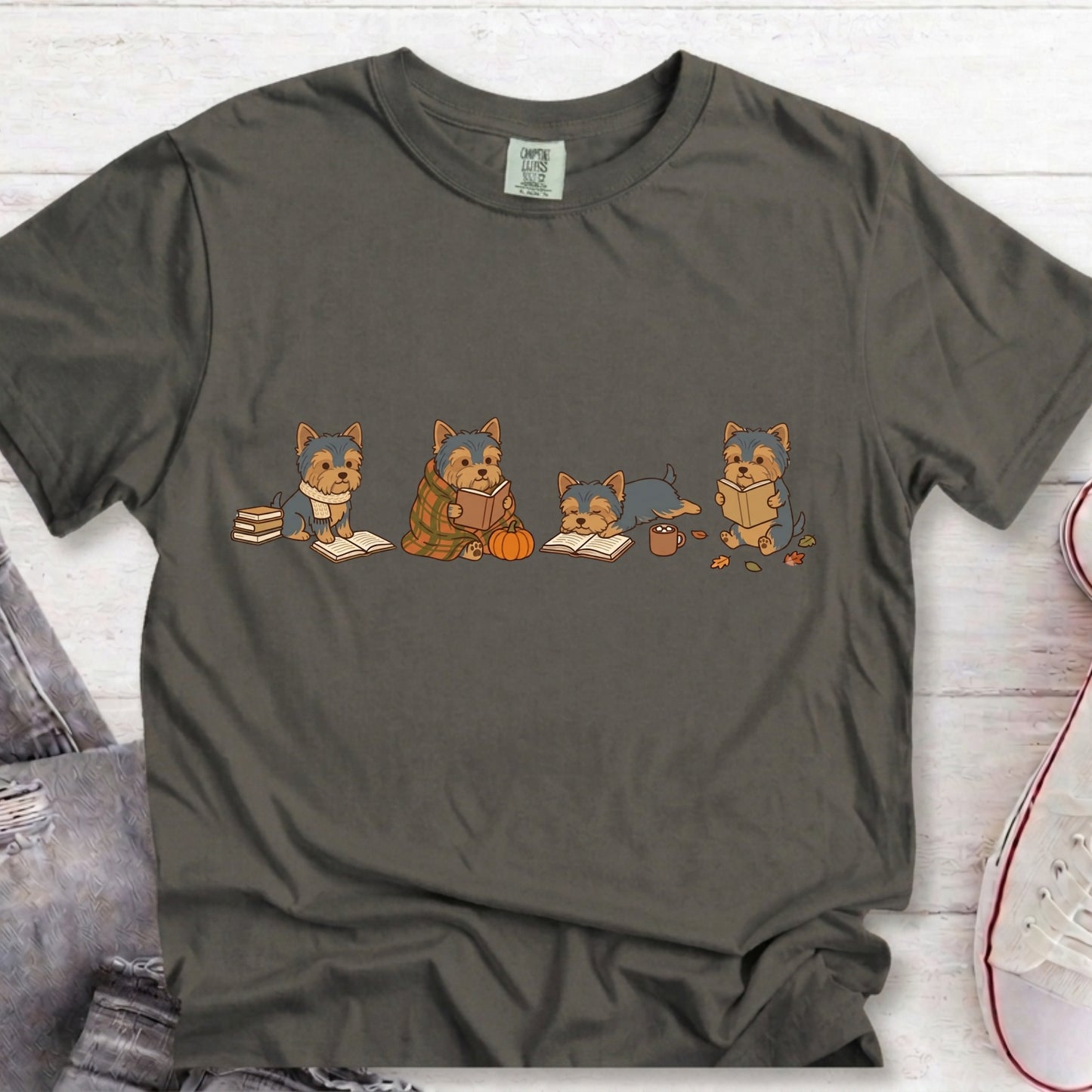A dark gray crewneck t-shirt featuring a cartoon graphic of four dogs reading books, laid flat on a white wood background with jeans and a sneaker partially visible.