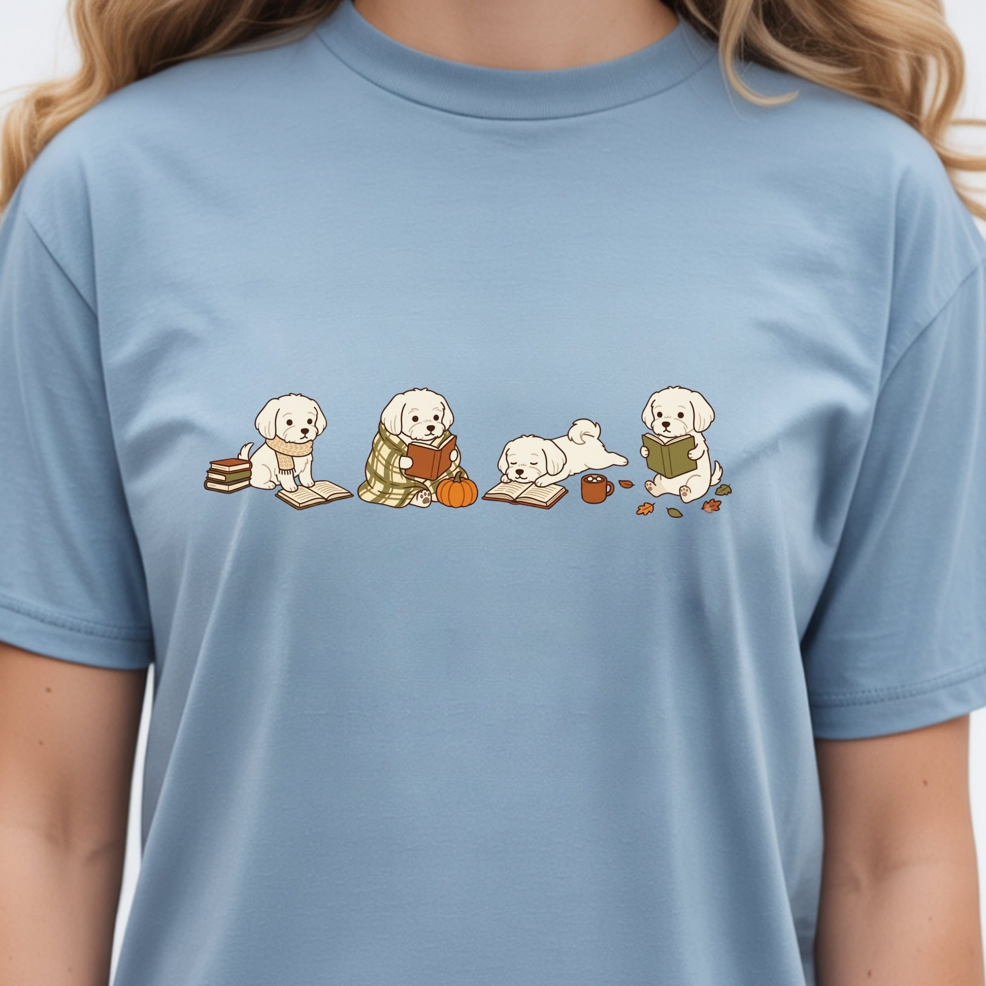 A light blue crewneck t-shirt featuring a graphic of four cute cartoon puppy dogs in autumn outfits (reading books, holding an acorn, lying down, and sitting up) worn by a model.